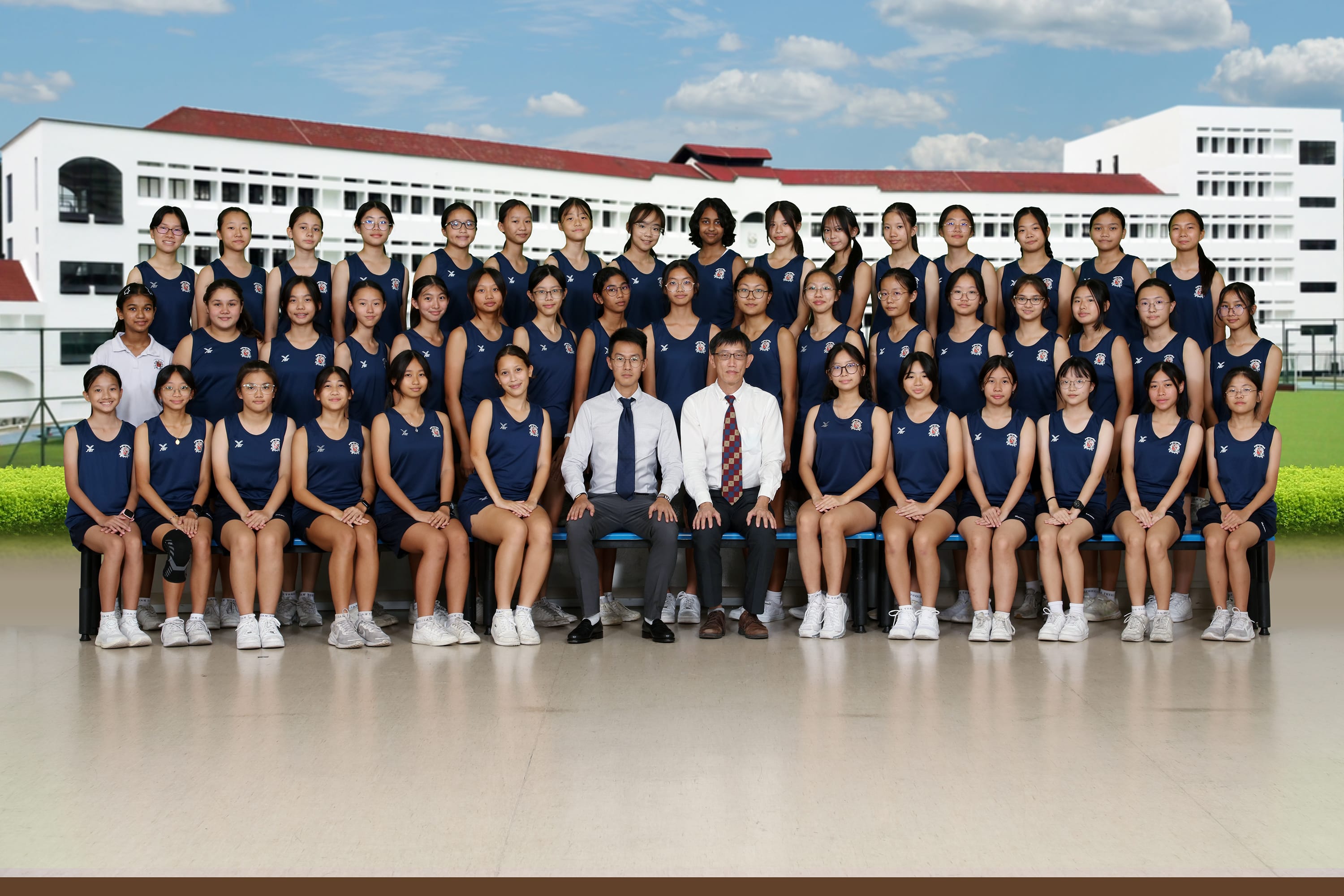 Large group of girls in navy sports uniforms posing with two male teachers in front of a white school building.