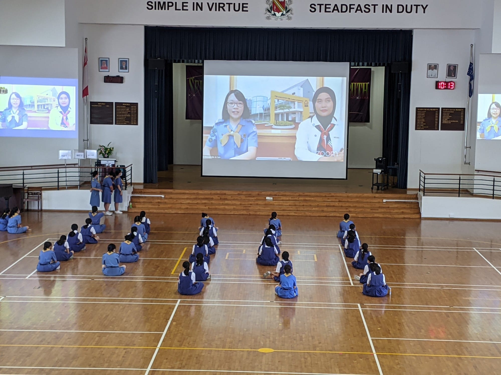 Students in blue uniforms sit spaced apart on a hall floor watching a Zoom call projected on a large screen.