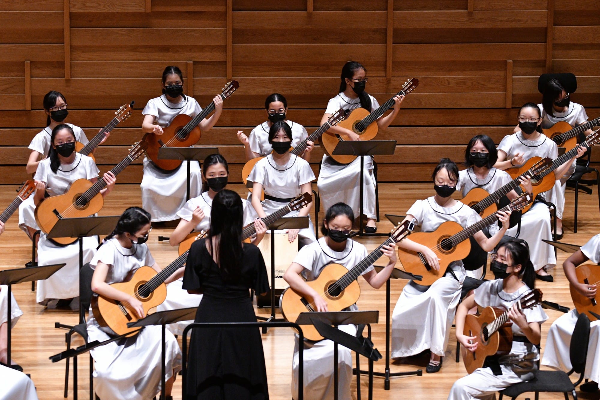Young girls in white dresses and black masks play classical guitars on stage, led by a conductor in black.