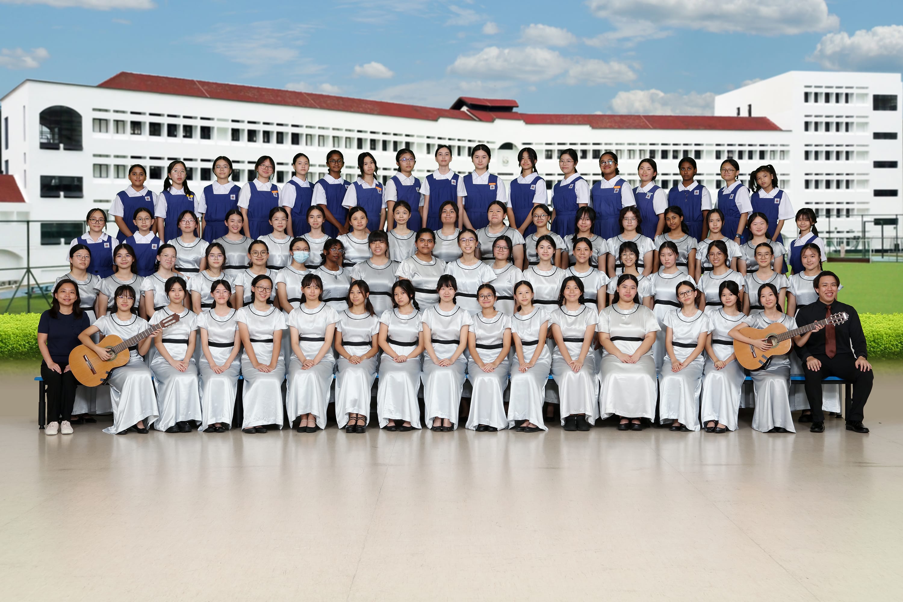 Large group of school girls in uniform pose in rows, with two guitar players on each side, in front of a school building.