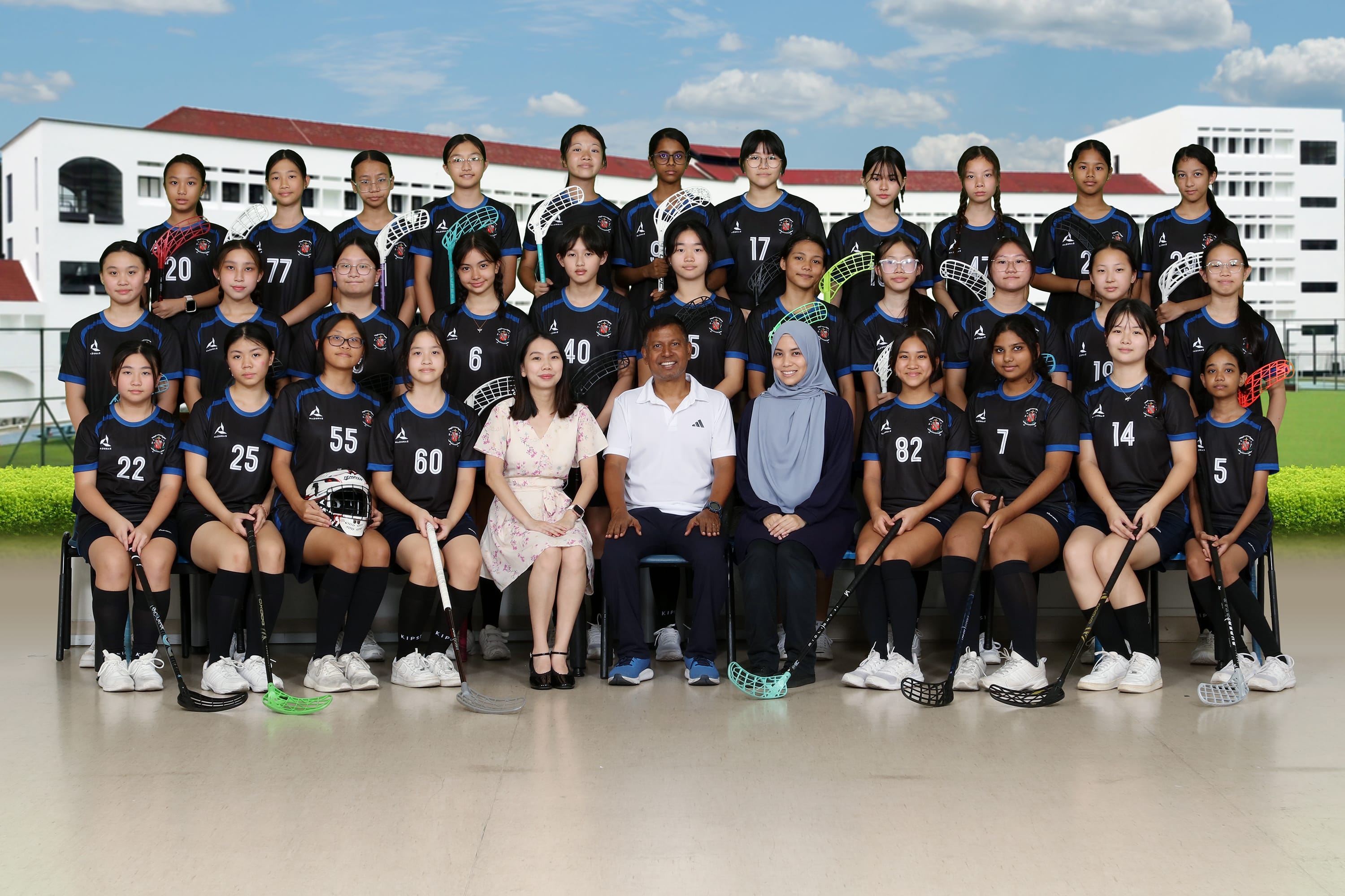 A girls floorball team in black jerseys poses with coaches in front of a school building.
