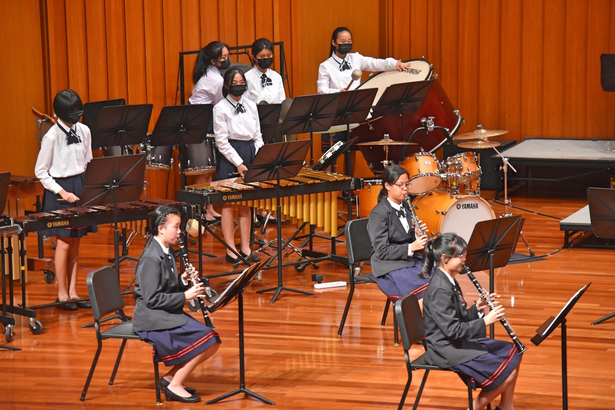 Student musicians in uniforms perform on stage with clarinets, xylophones, and Yamaha drums.