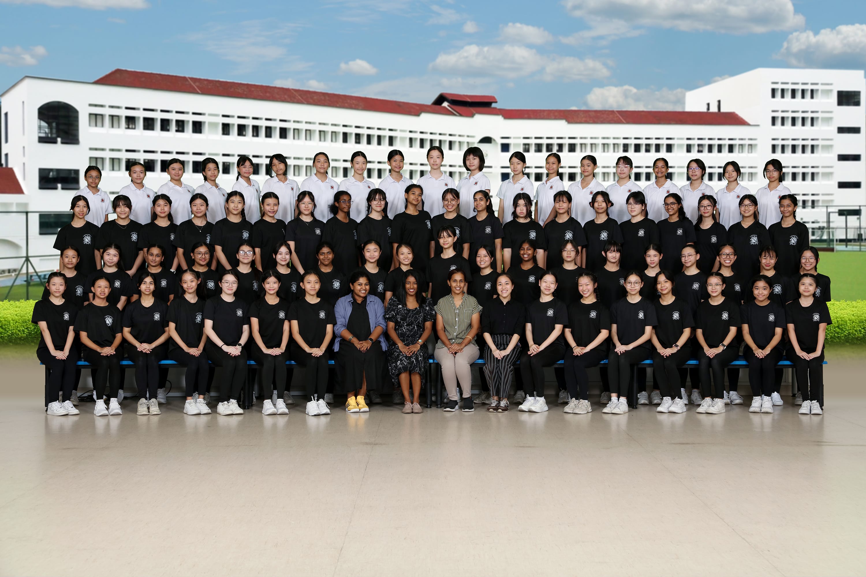 Large group of school girls in black uniforms posed in rows with teachers in front of a white school building.