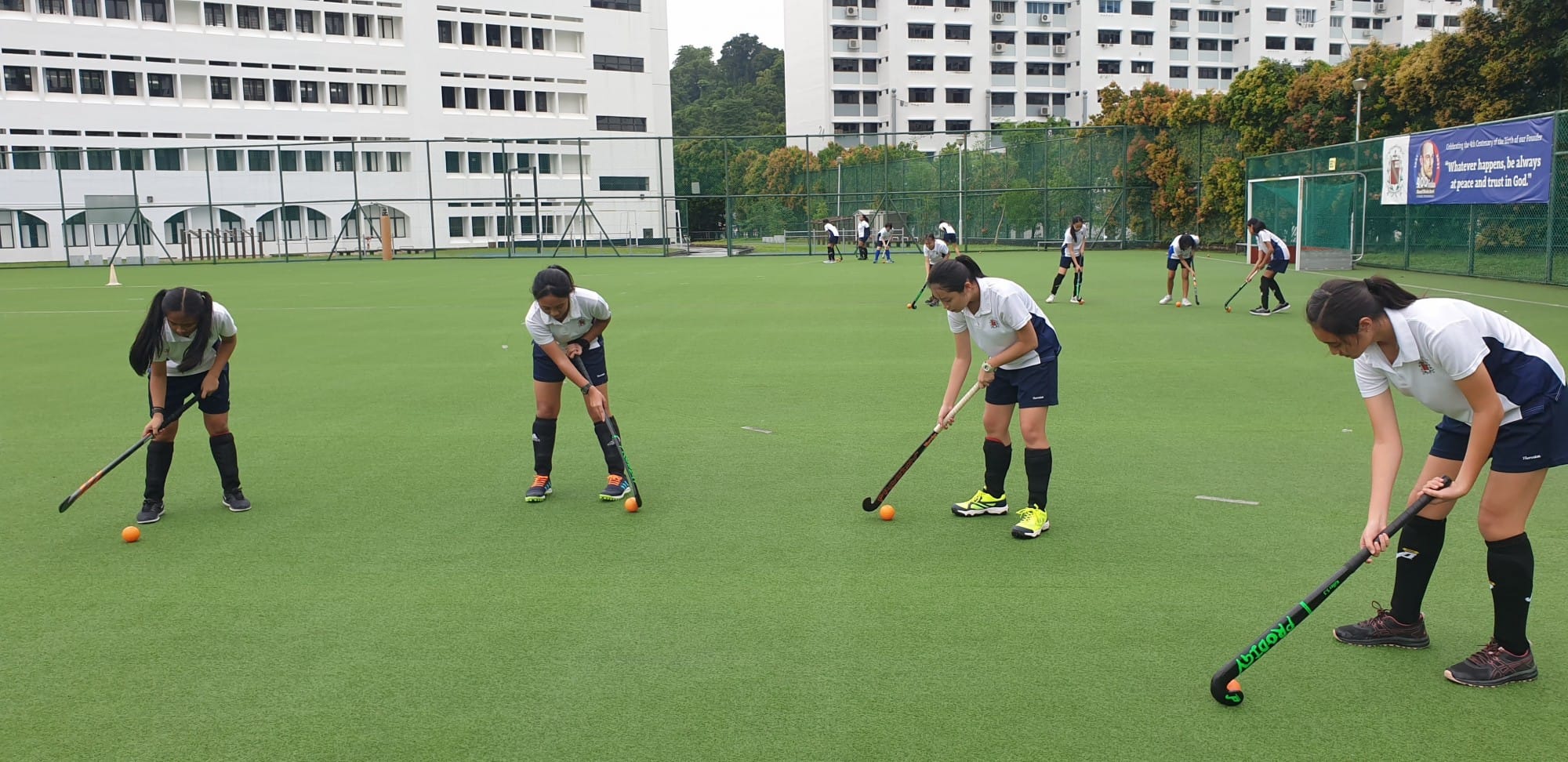 Girls in white and navy uniforms practice field hockey with sticks and orange balls on a green turf court.