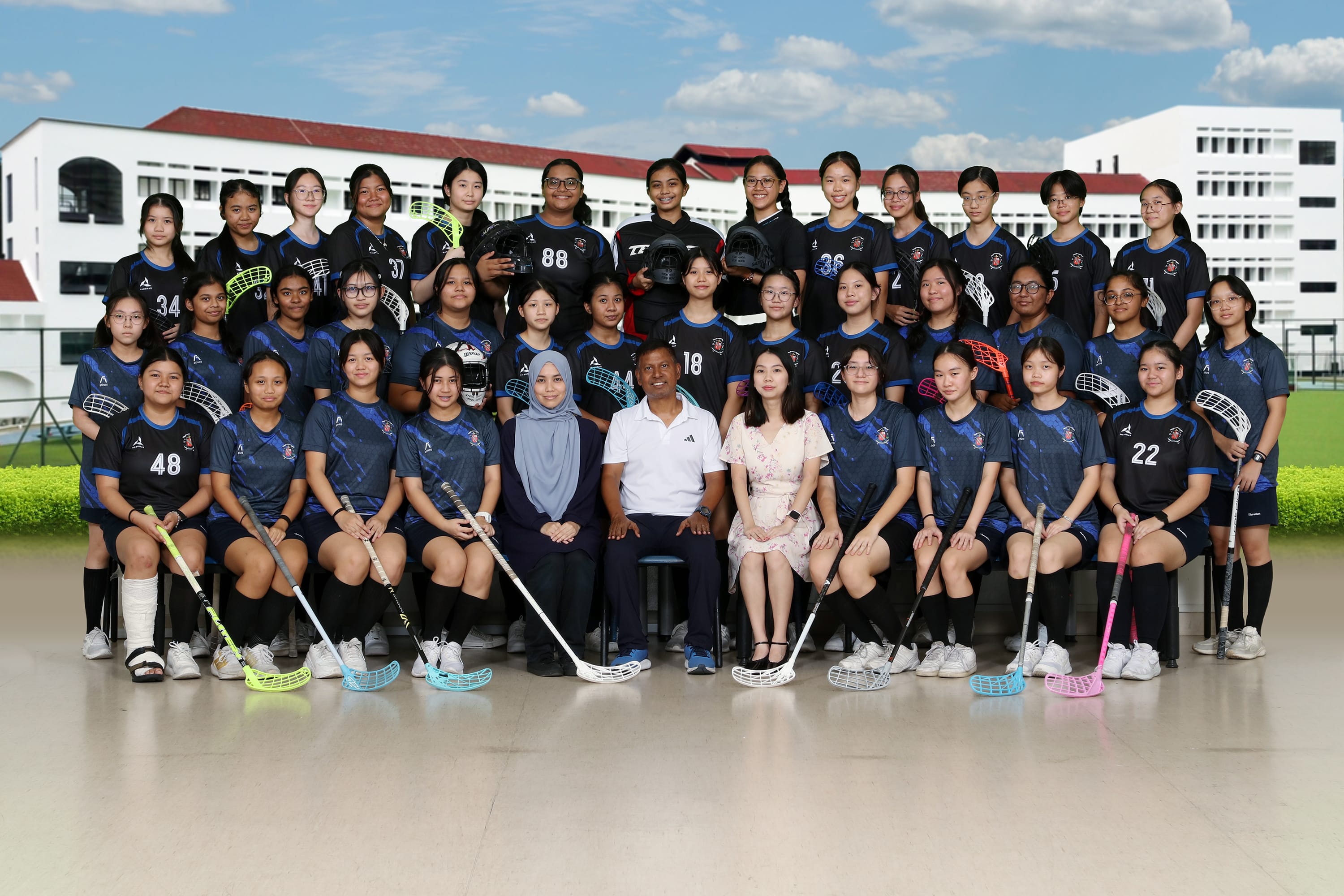 A girls floorball team in dark blue jerseys poses with sticks and helmets alongside two coaches outdoors.