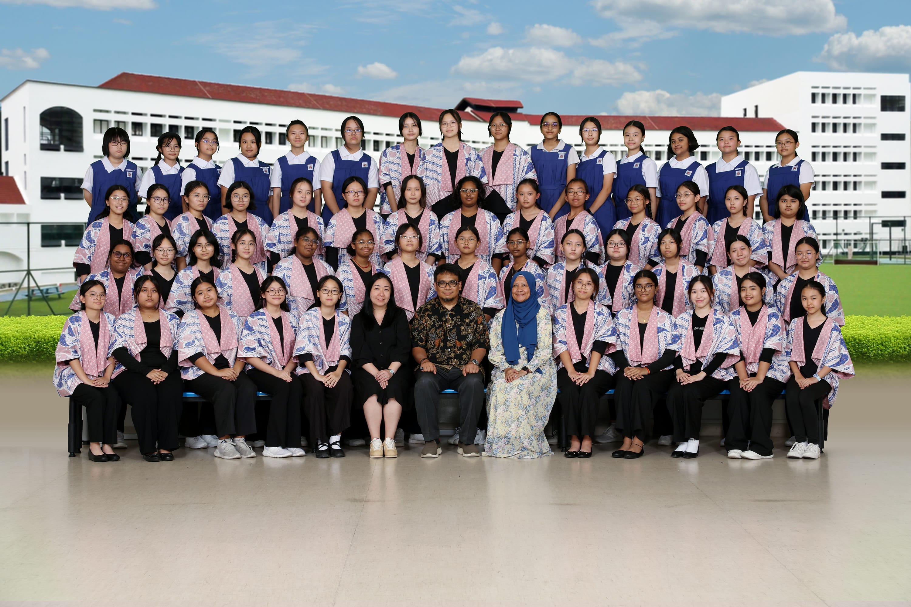 Large group of students in blue uniforms or black outfits with pink sashes posing with three teachers outdoors.