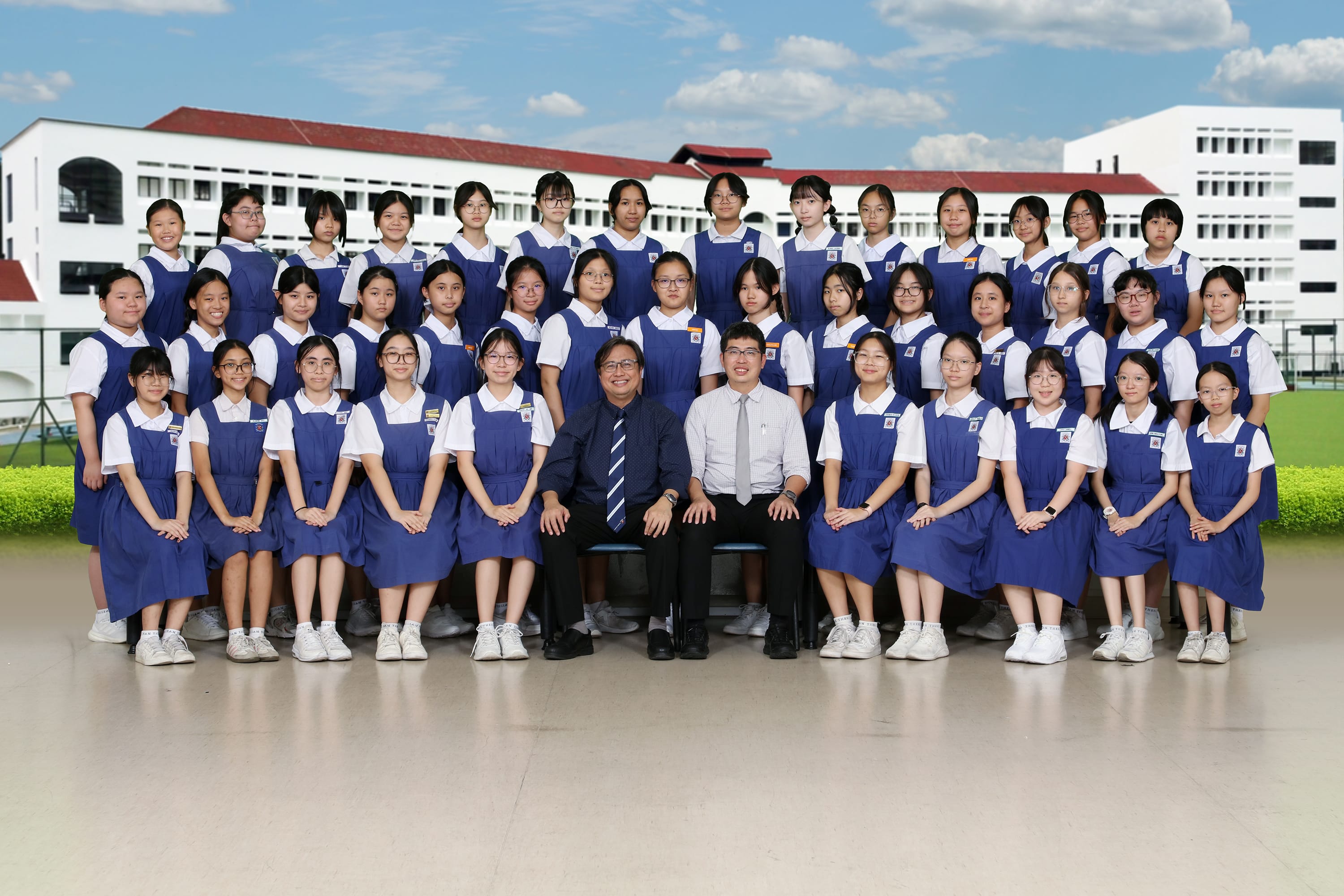 Large group of schoolgirls in blue uniforms posing with two male teachers in front of a school building.