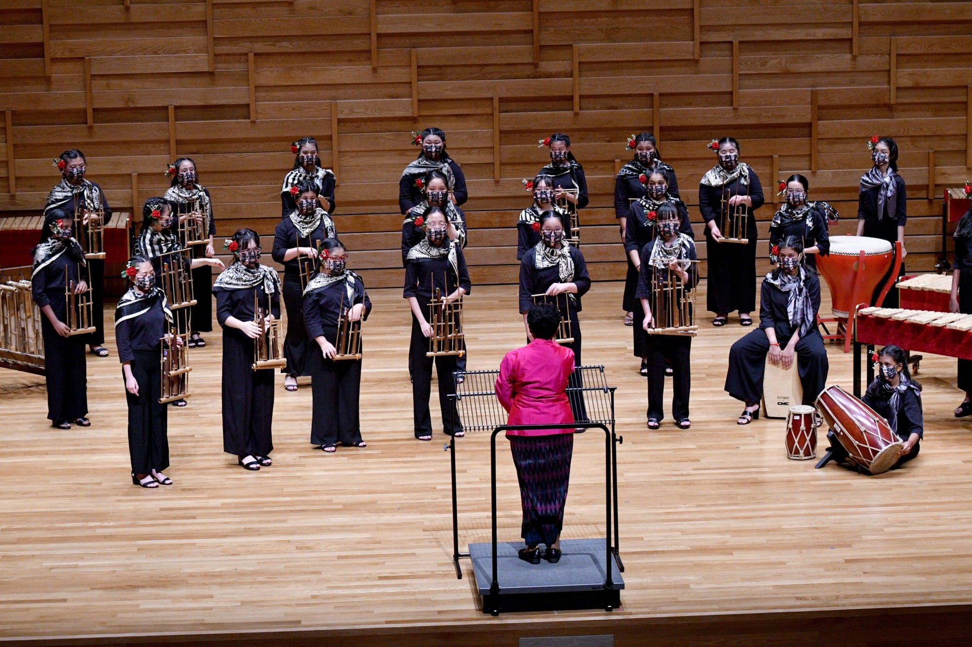 A conductor in red leads masked performers in black holding angklung instruments on a wooden concert stage.