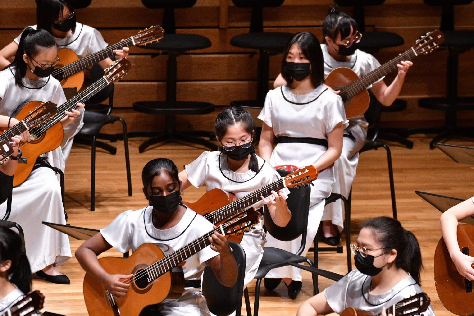 Young girls in white dresses and black masks play classical guitars on a wooden concert stage.