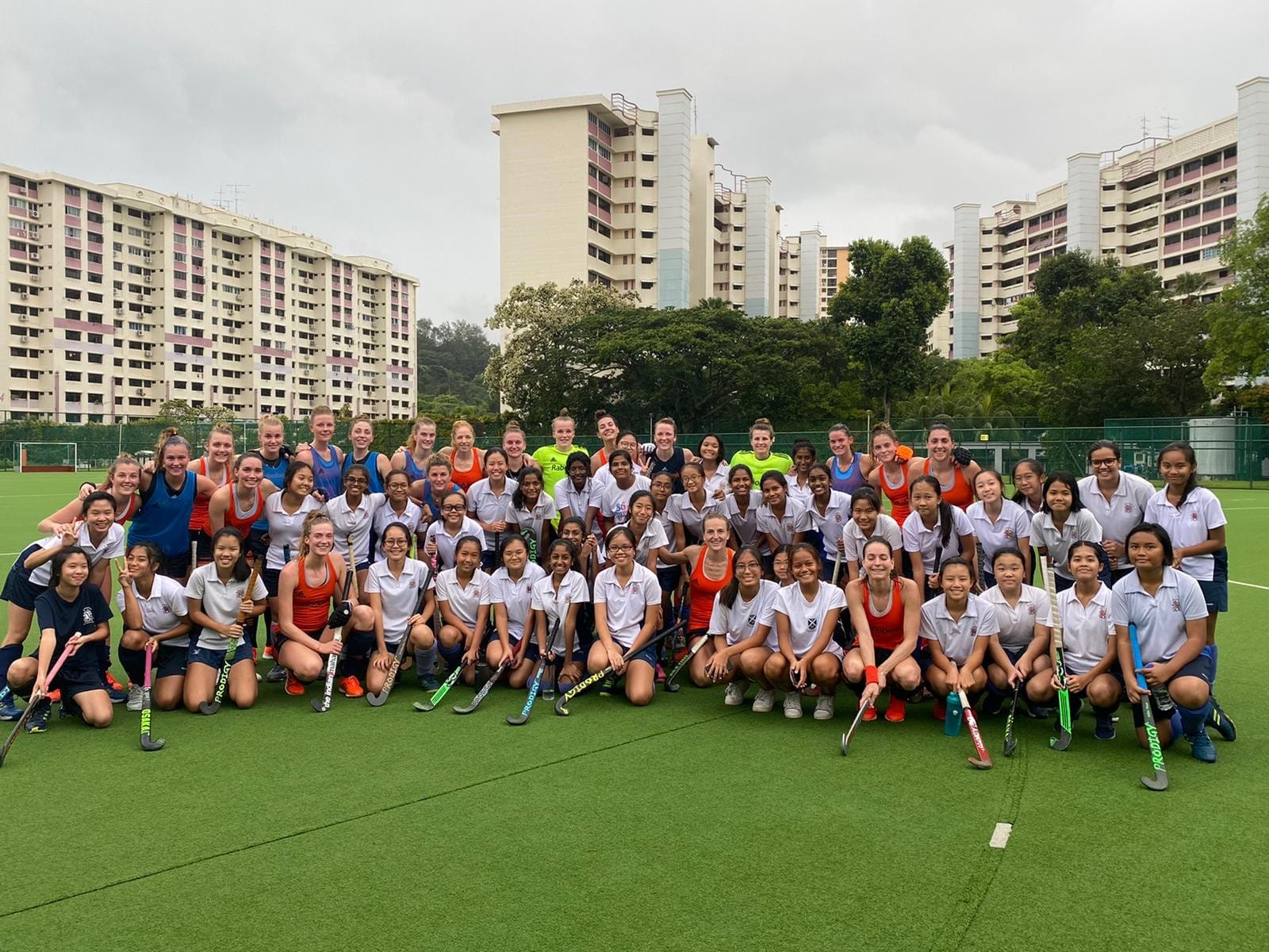 Large group of girls and women holding field hockey sticks posing on a green turf pitch with tall buildings behind.