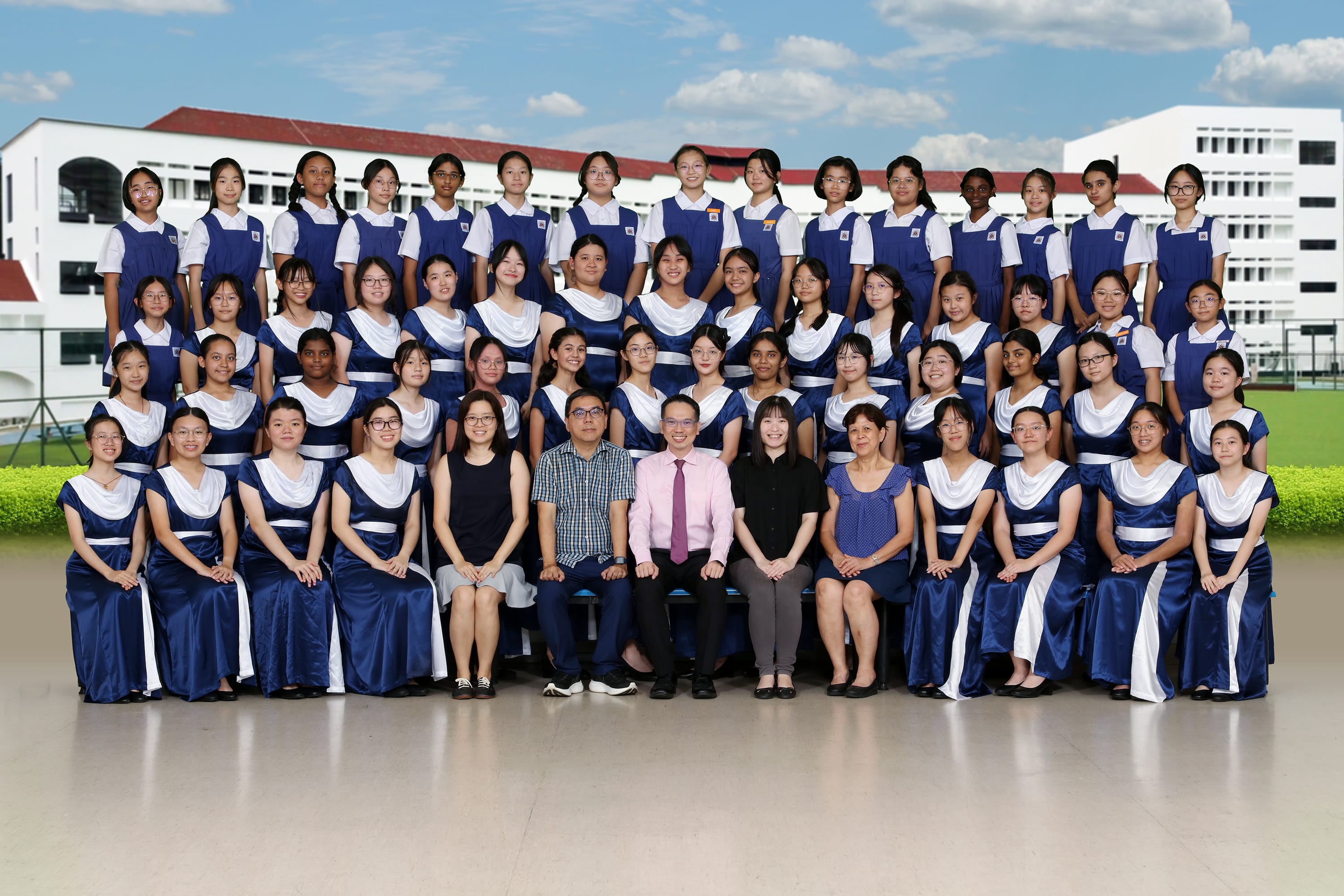 Large group of female students in blue and white uniforms posed in rows with several seated adults in front of a school building.