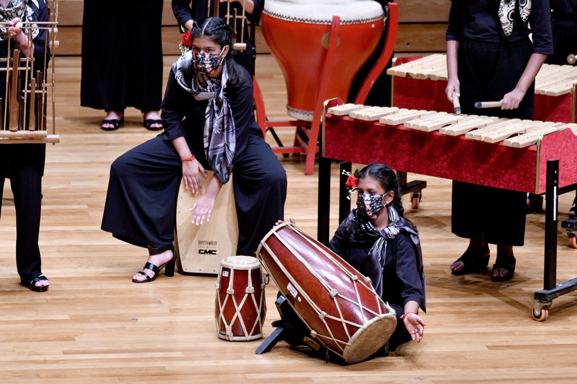 Two masked girls in black perform with drums and a cajon on a wooden stage surrounded by a marimba and timpani.