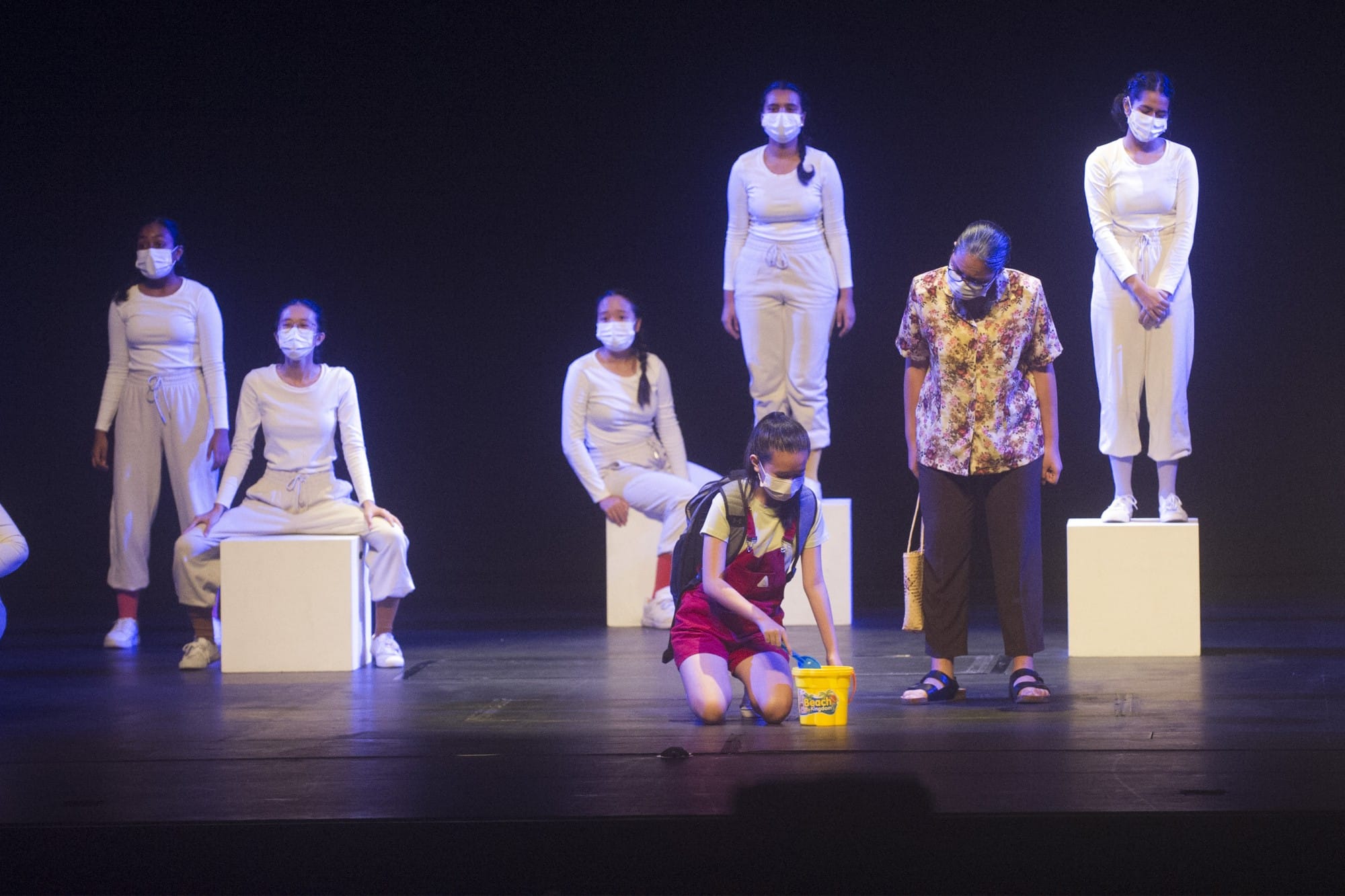 Stage performers wearing masks, most in white outfits, with a child kneeling holding a yellow bucket at center.