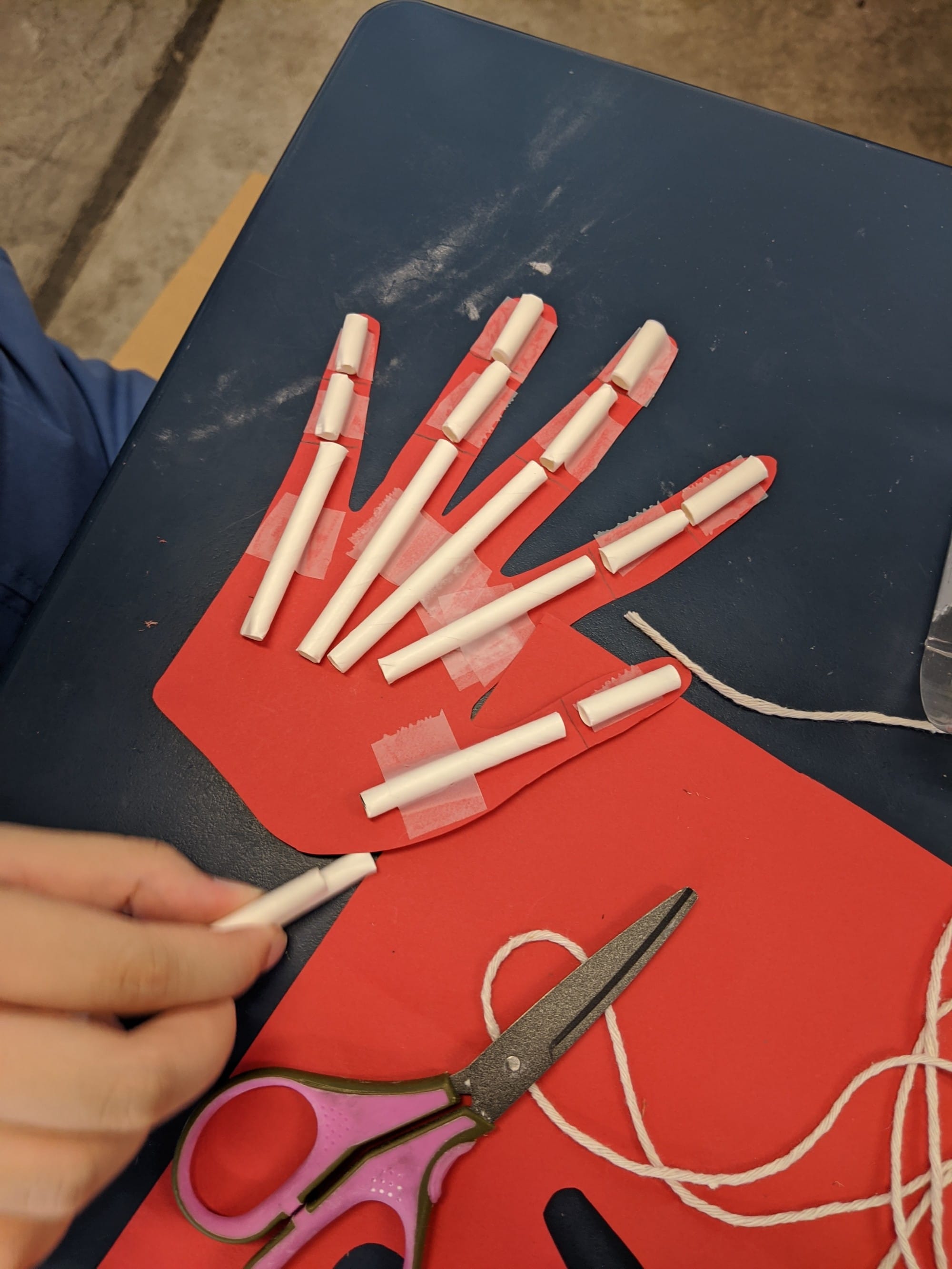 Red paper hand cutout with paper tube finger joints taped on, string and scissors nearby on a blue desk.