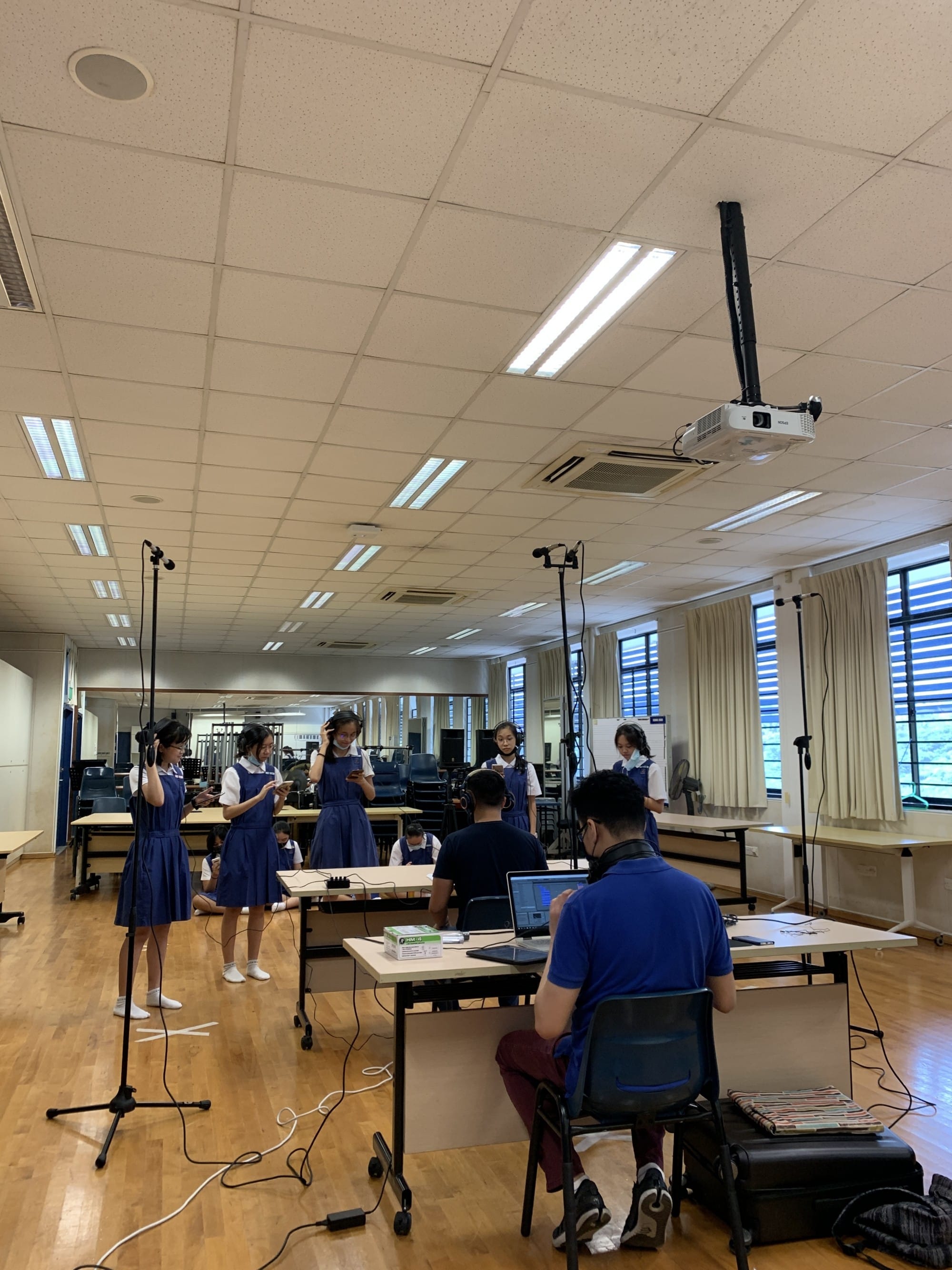Students in blue uniforms stand near microphone stands in a classroom while a person sits at a laptop with headphones.