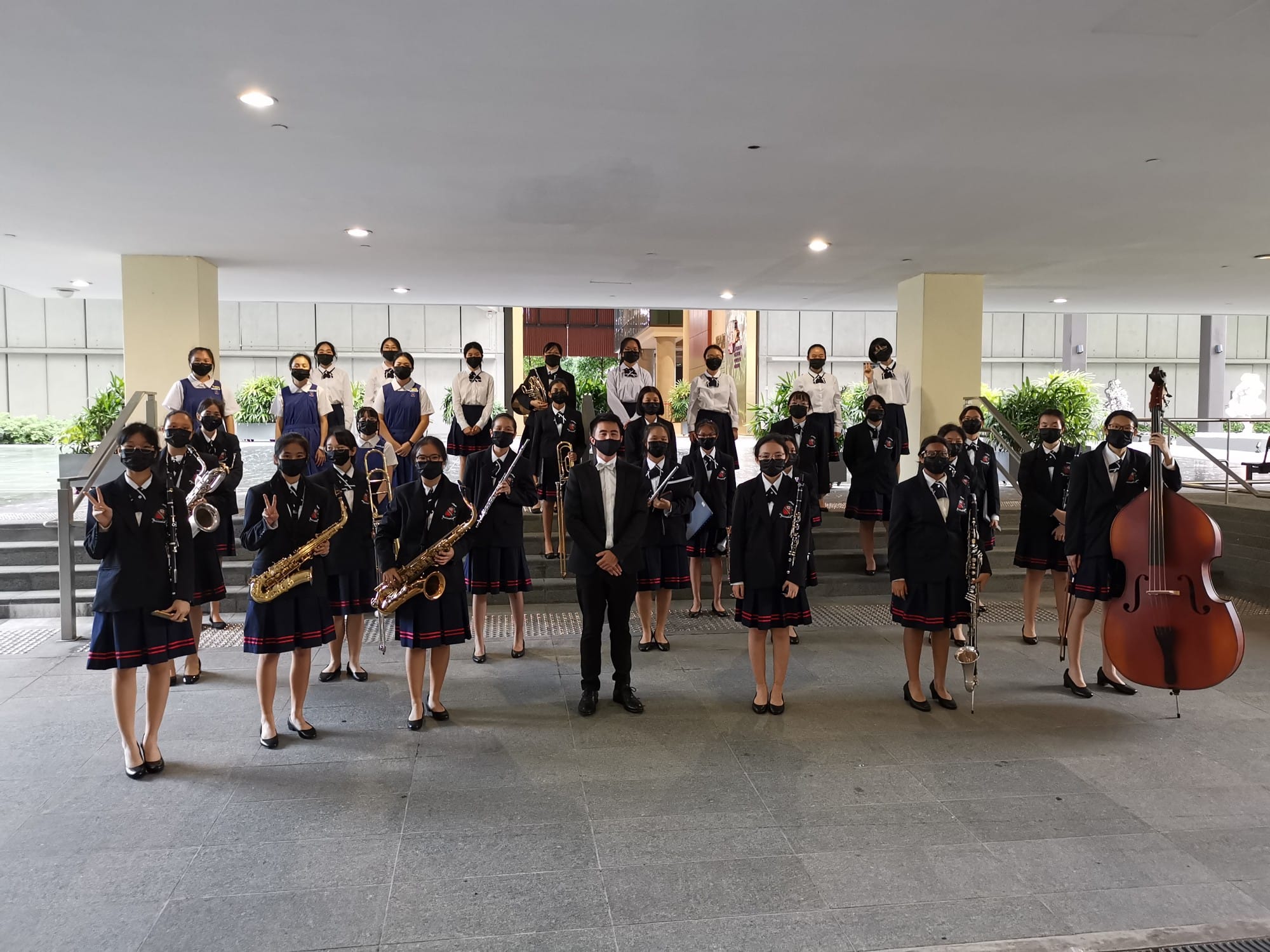 A school band in uniforms and masks poses indoors holding saxophones, clarinets, a double bass, and other instruments.