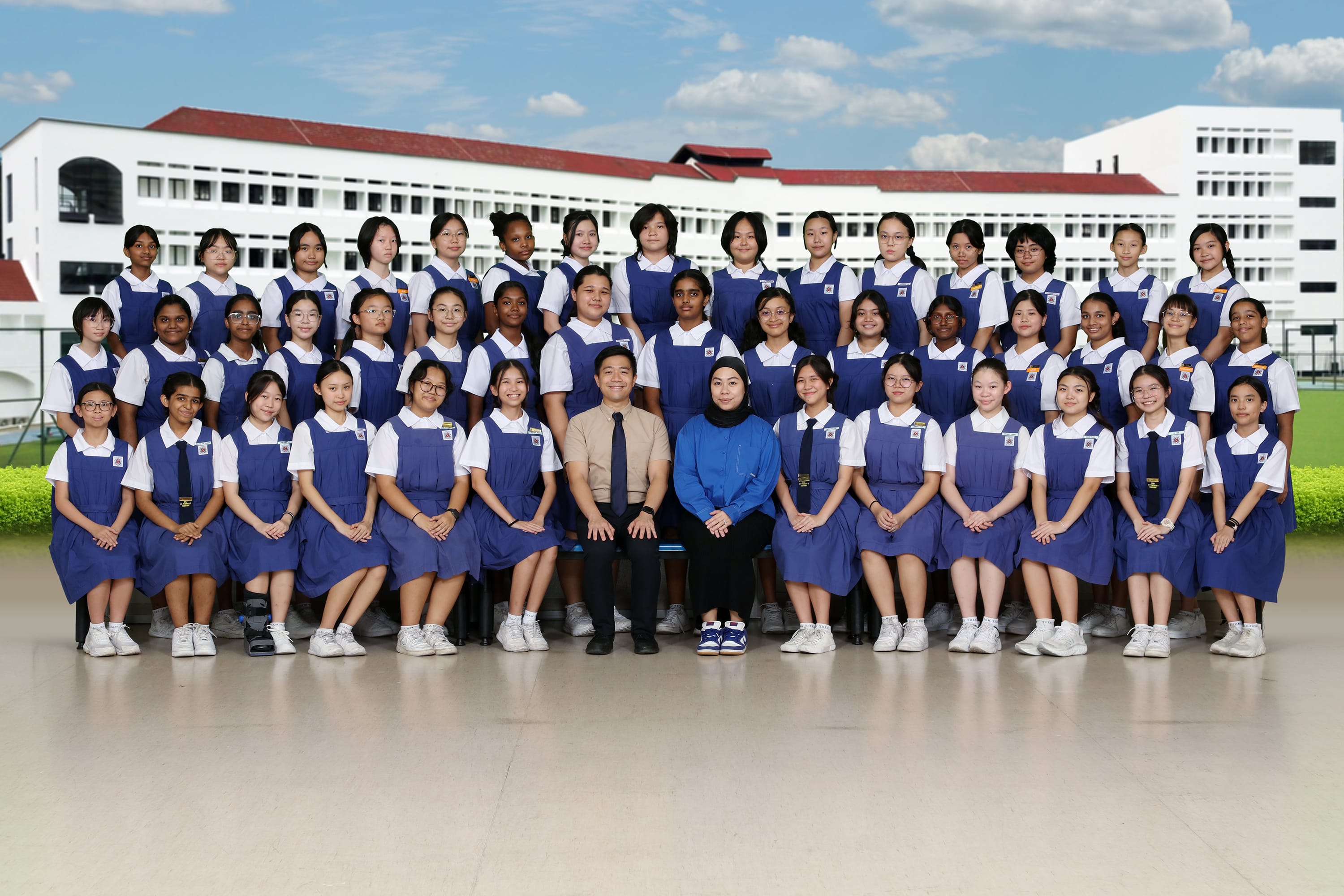 Large group of schoolgirls in blue pinafore uniforms posed in rows with two teachers seated in the center.