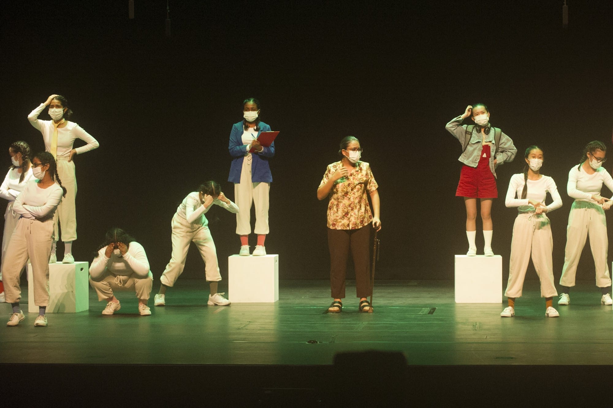 A group of masked performers on a dark stage, some standing on white boxes, in various dramatic poses.