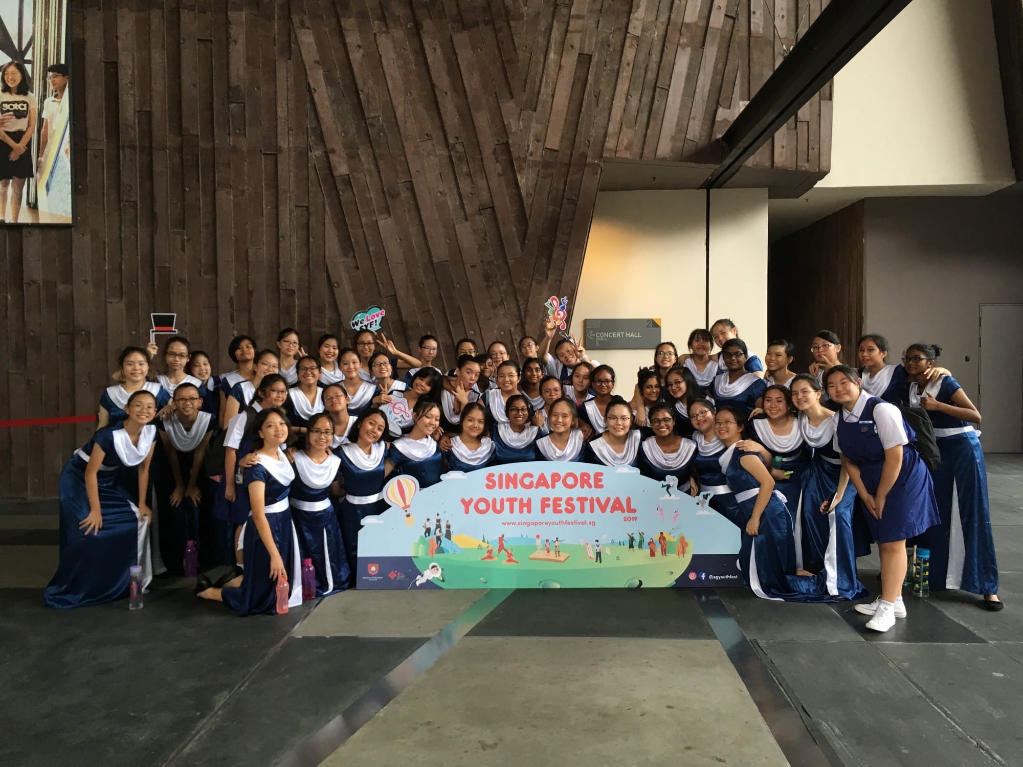 A large group of young women in blue and white uniforms posing behind a Singapore Youth Festival 2019 banner.