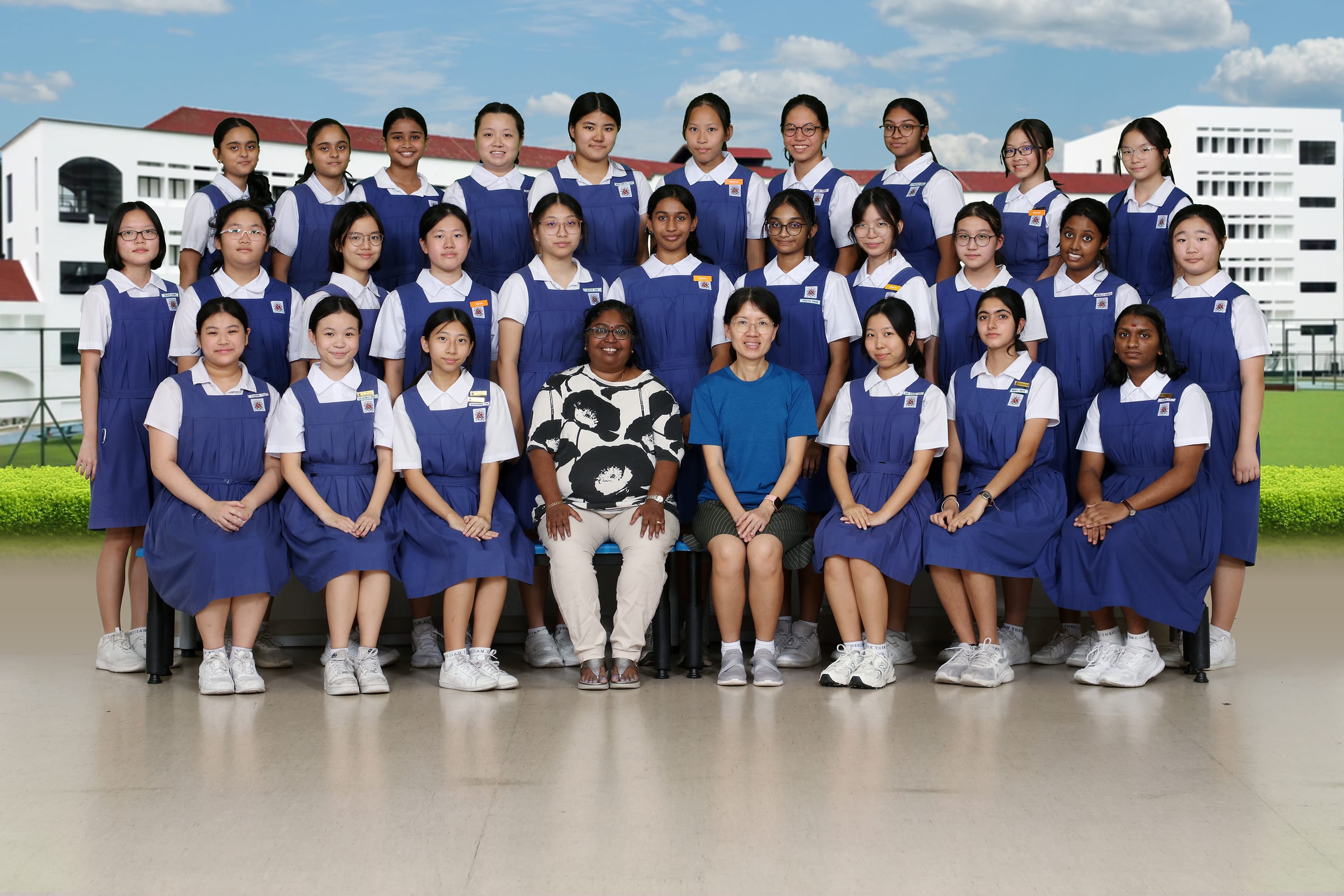 Group photo of schoolgirls in blue uniforms posed in three rows with two adult women seated in the center.