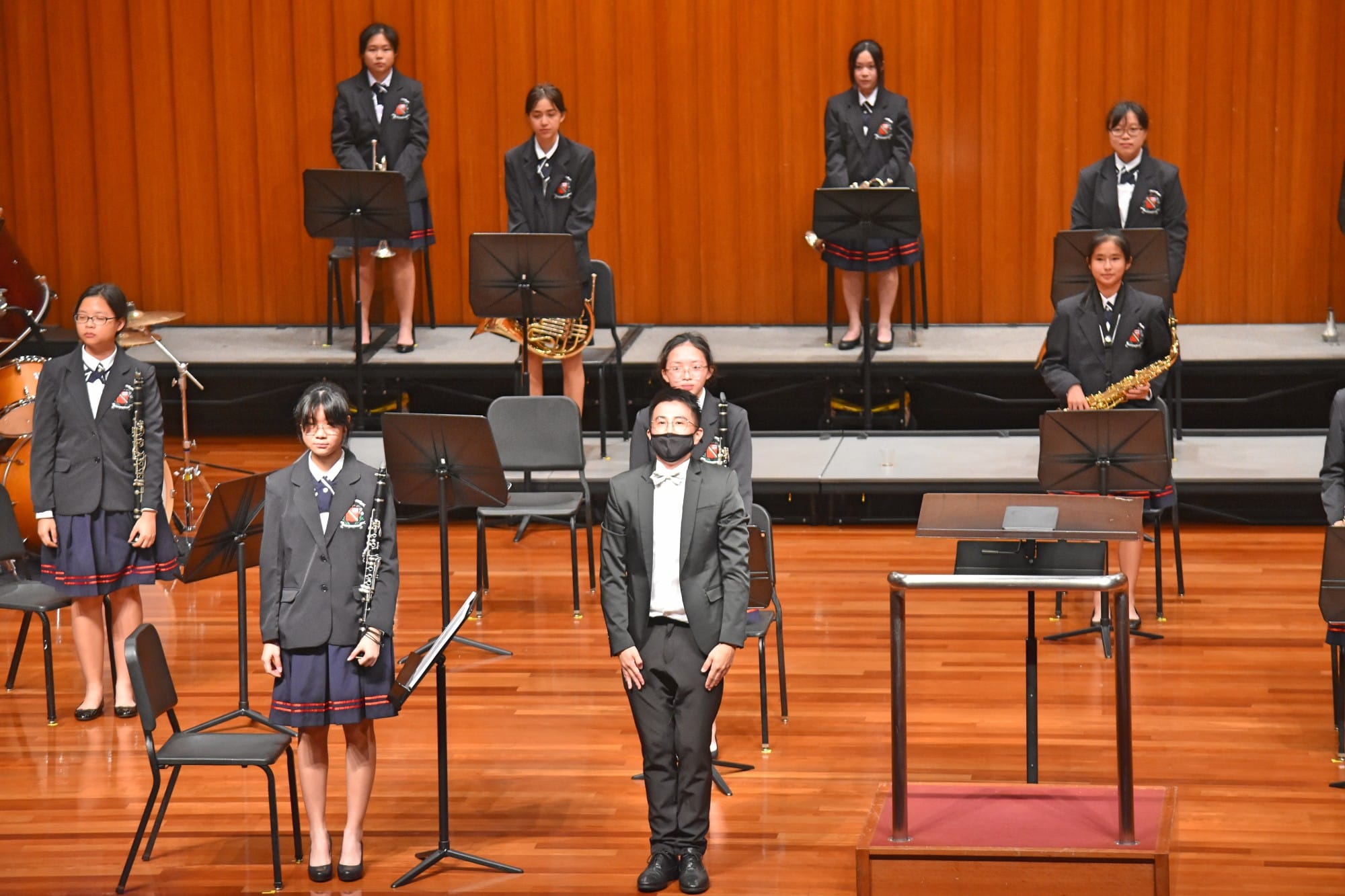 Student band members in dark uniforms stand on a wooden concert stage with music stands and instruments.
