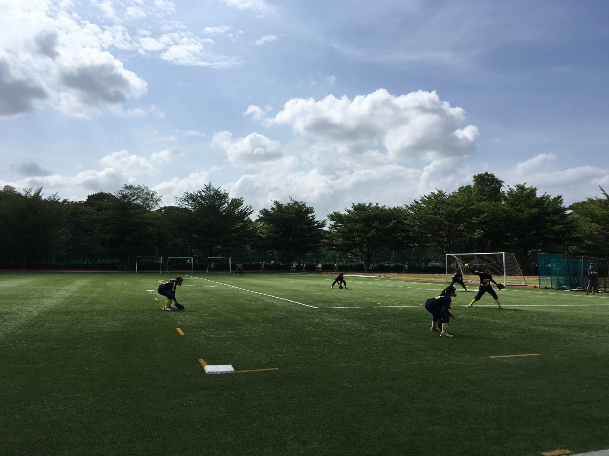 Players in navy uniforms practice softball on a green turf field surrounded by trees under a partly cloudy sky.