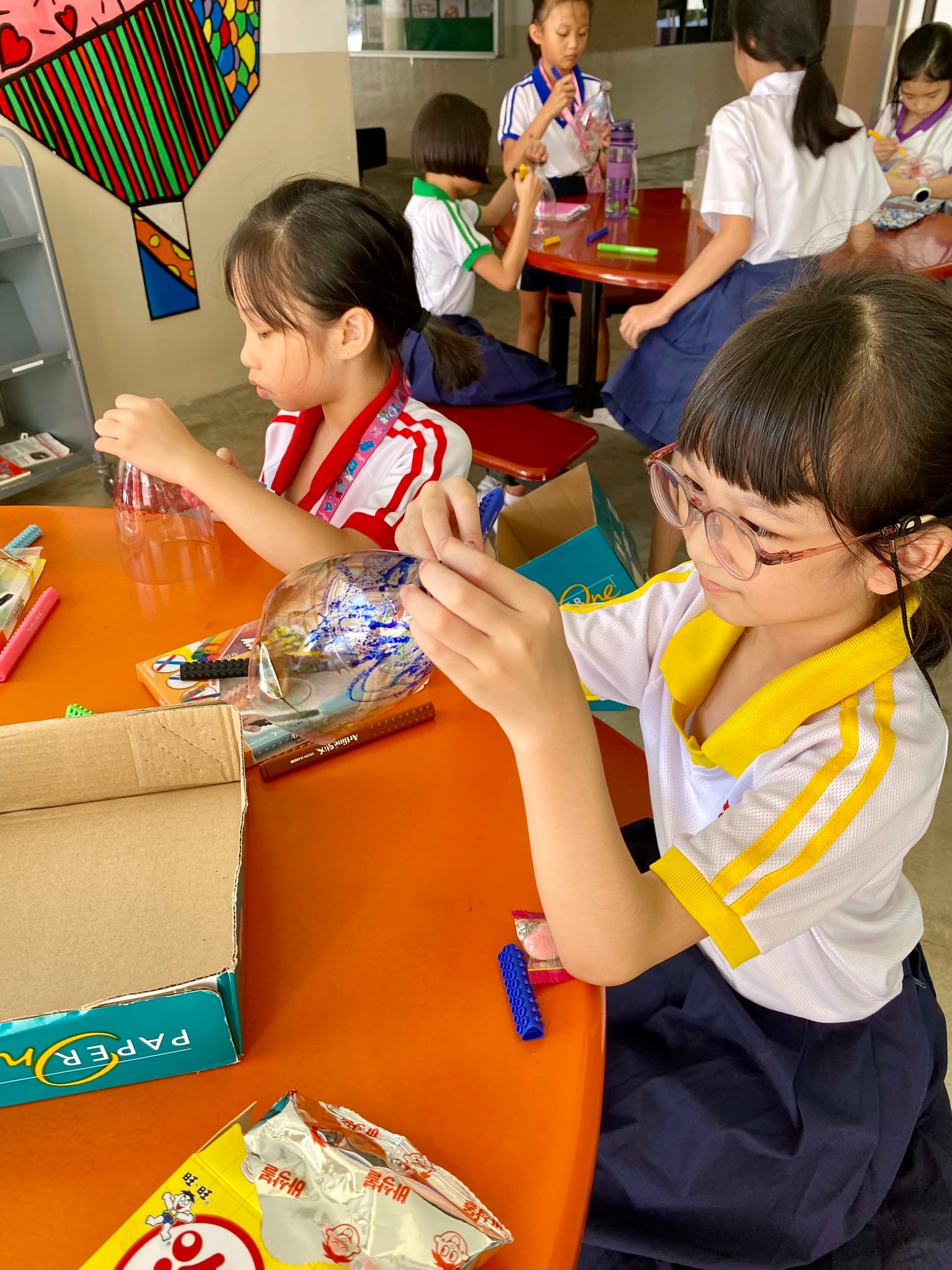 Young girls in school uniforms sit at an orange table, drawing on clear plastic objects with markers.