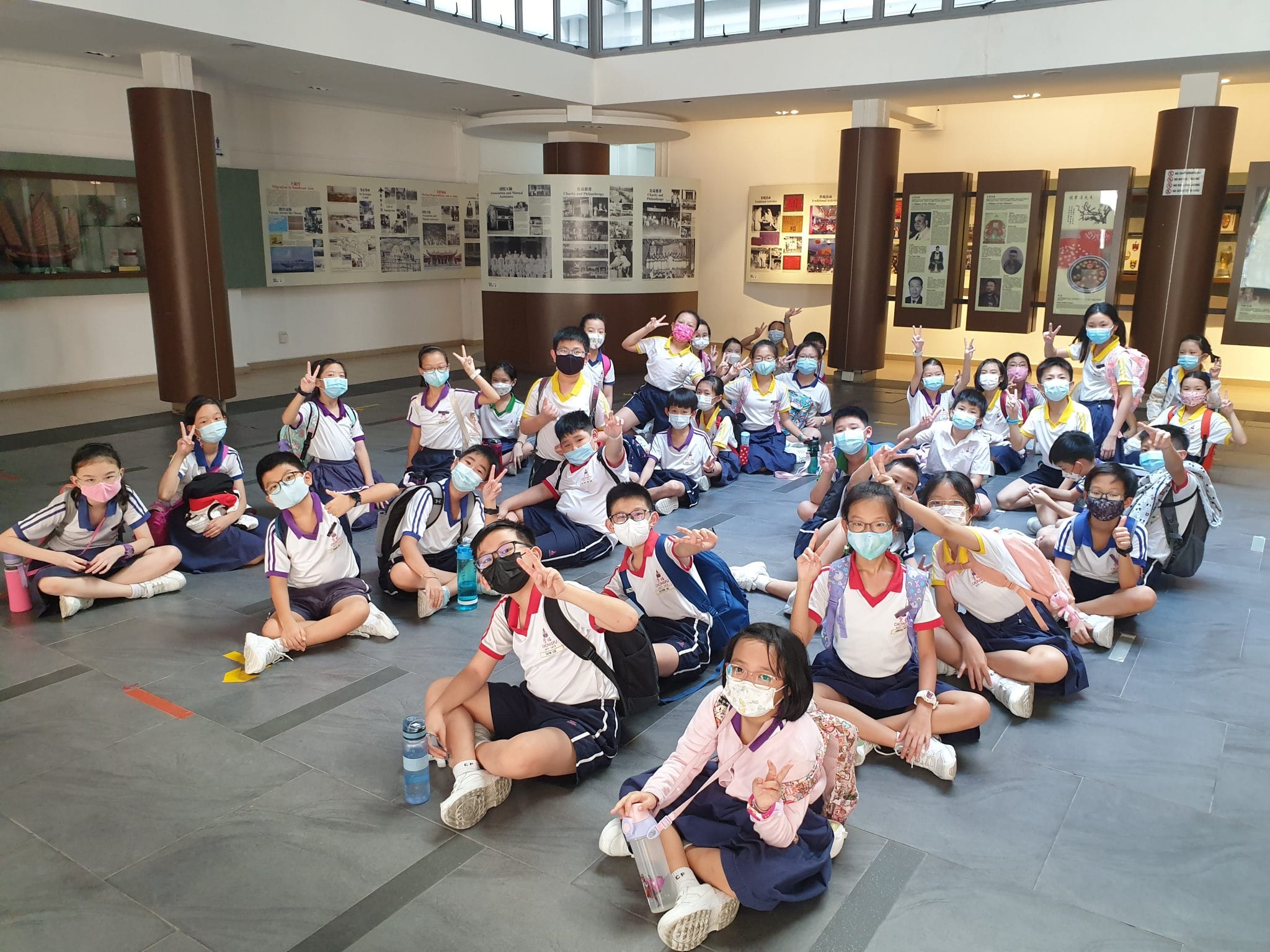 A large group of masked schoolchildren in uniforms sit on a museum floor, many making peace signs.