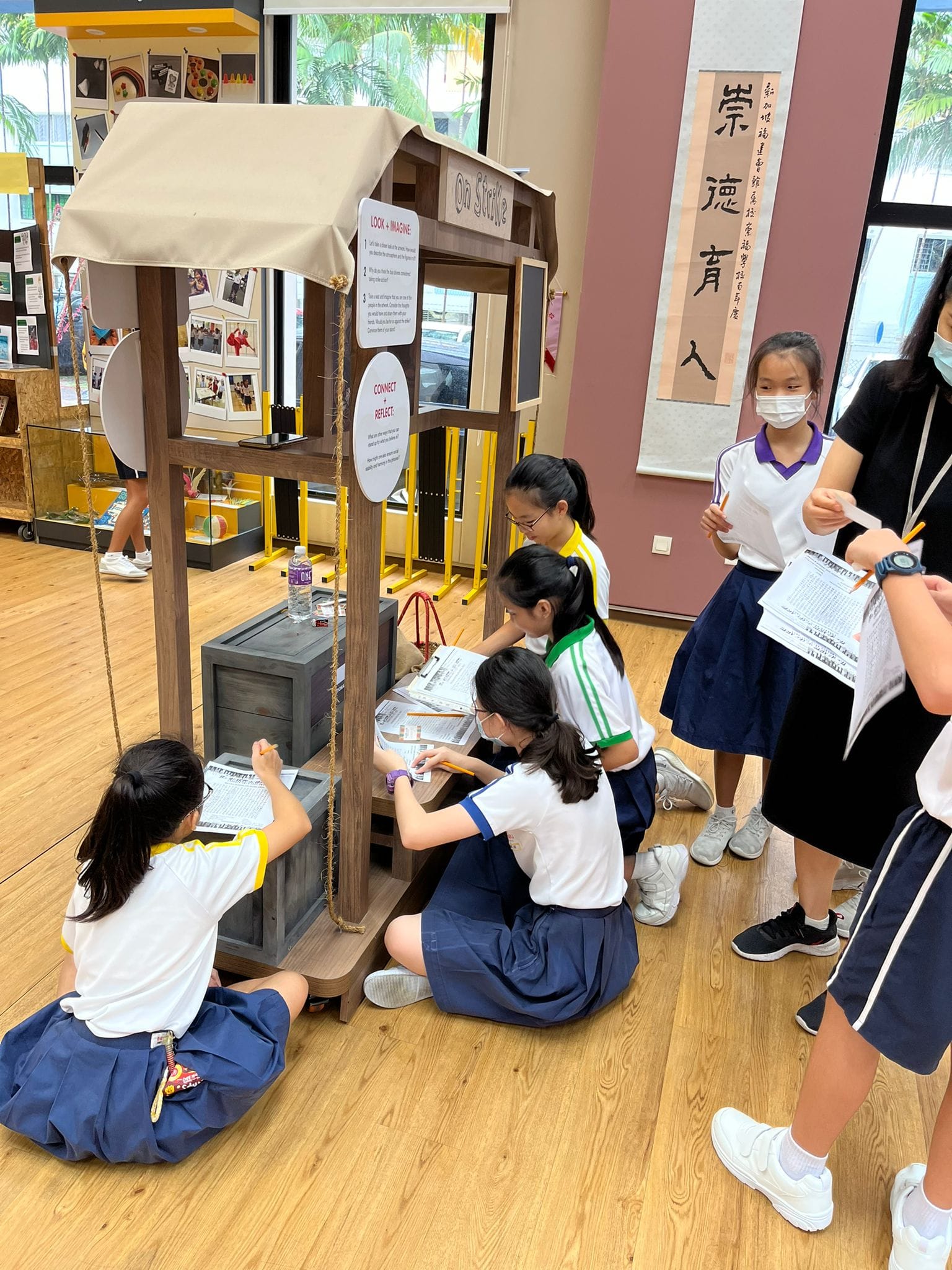 Several school girls in uniforms gather around a wooden booth labeled "On Strike" inside a hall with Chinese calligraphy.