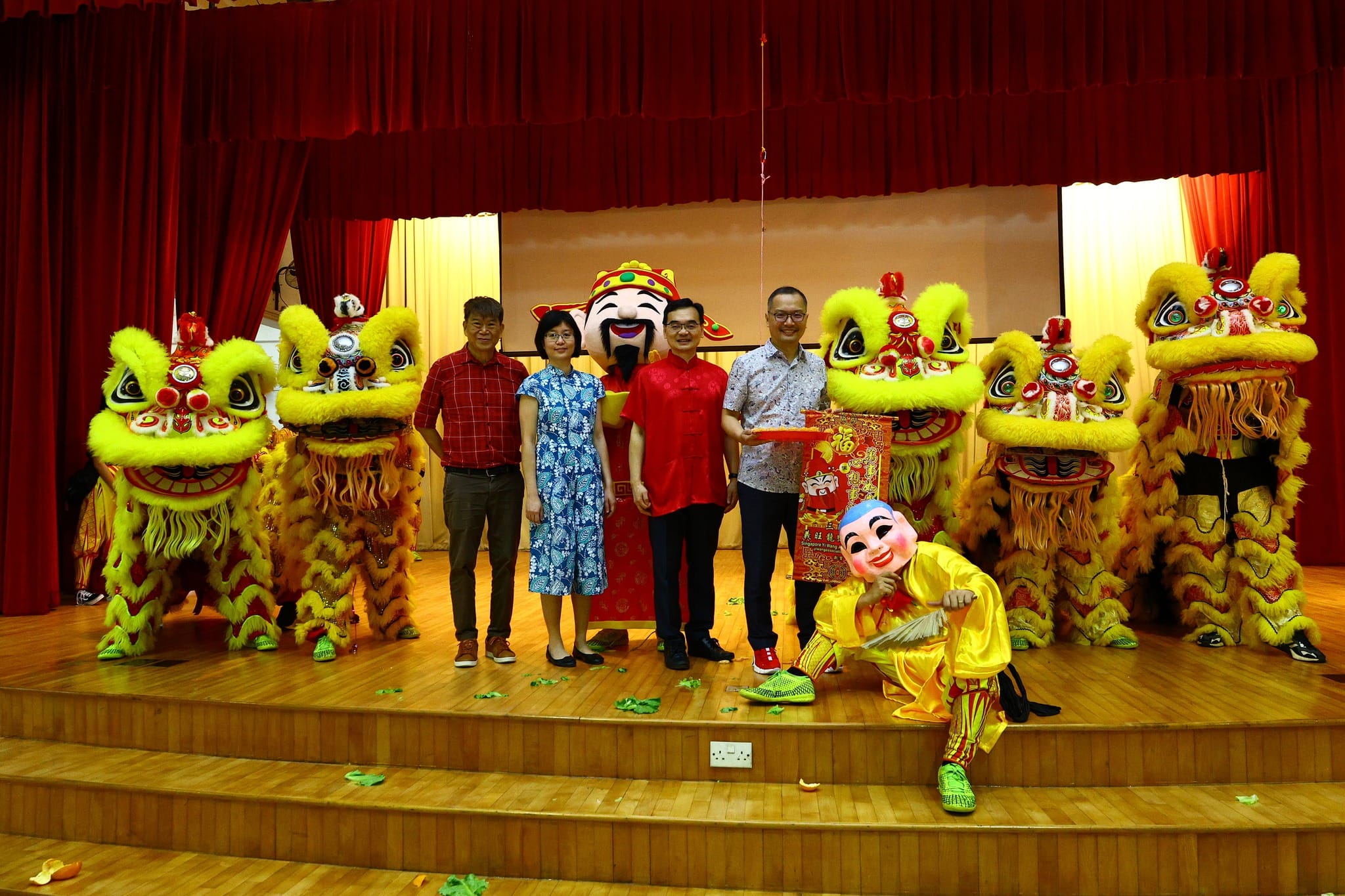 Four people pose on stage with yellow lion dance costumes and performers in colorful Chinese New Year attire.