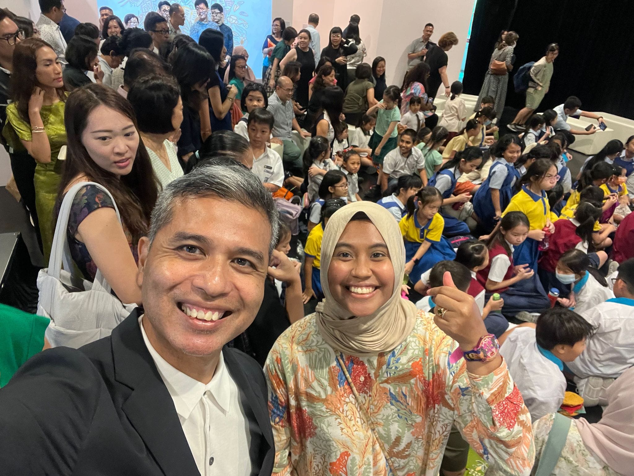 Two smiling adults take a selfie at a crowded indoor event with many children in school uniforms seated behind them.