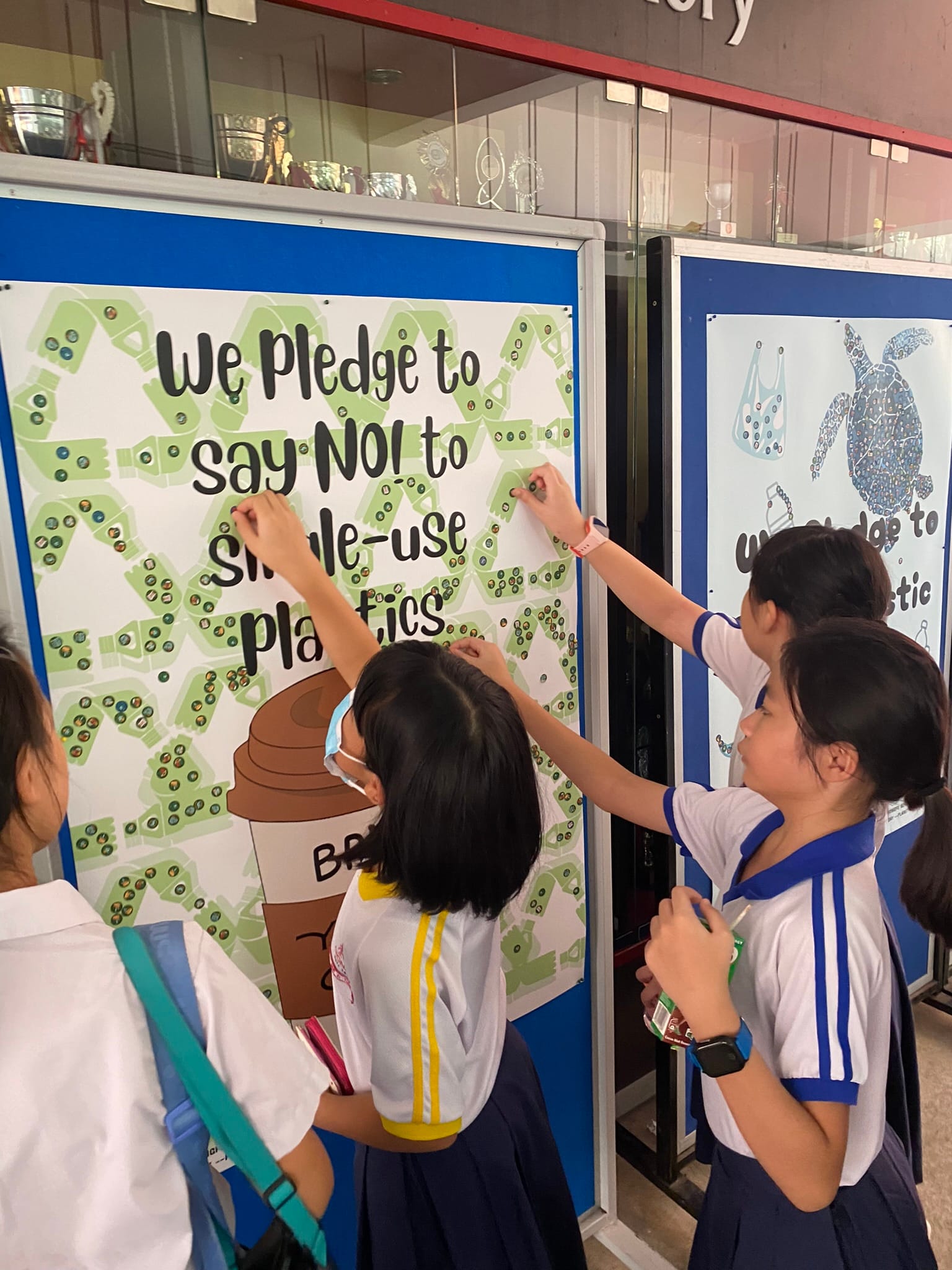 Students in school uniforms placing stickers on a pledge board reading "We Pledge to Say No to Single-use Plastics."