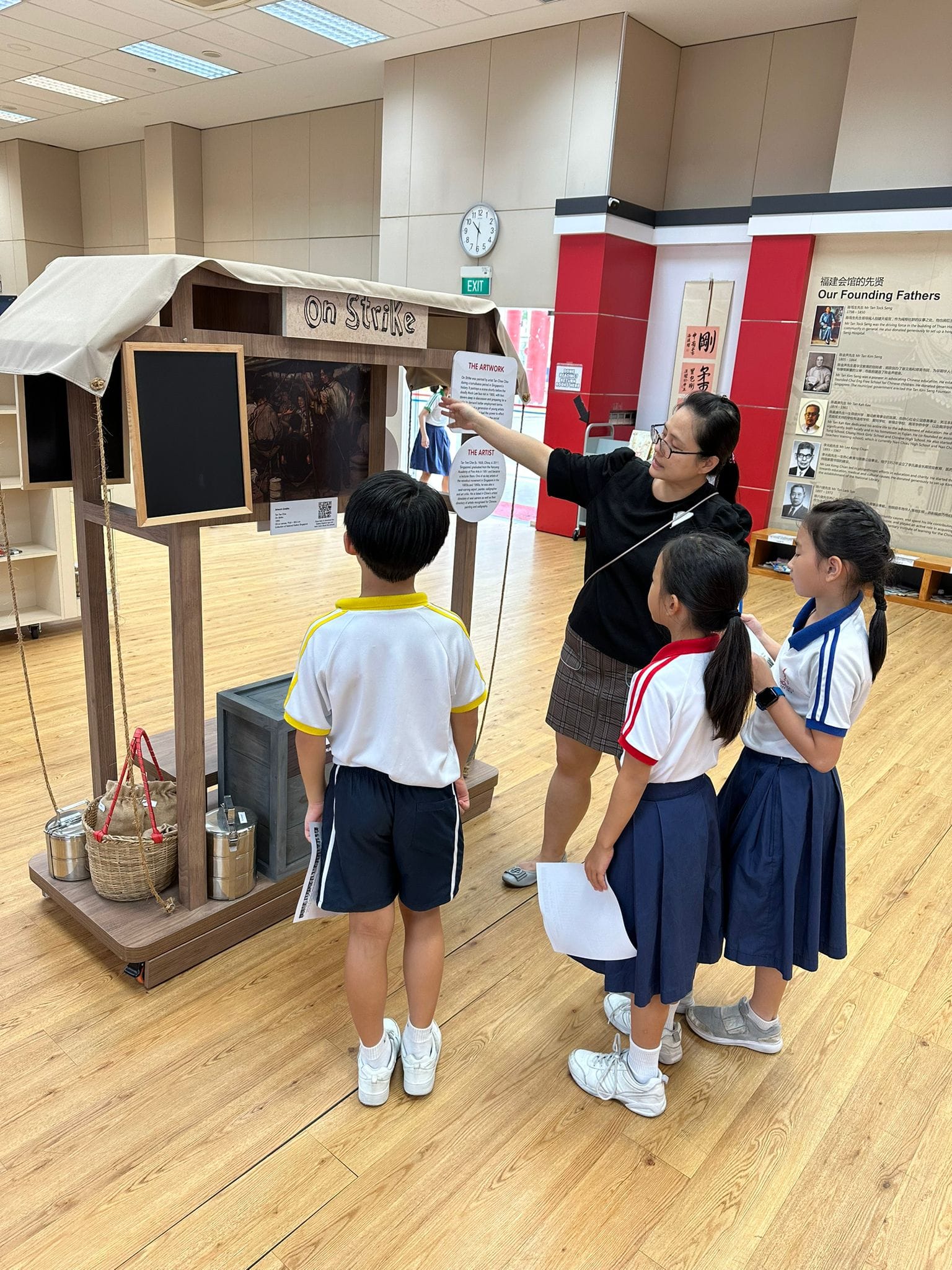 A teacher points to an "On Strike" exhibit display while three young students in uniforms look on in a museum.