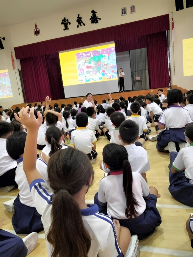 Students in white uniforms sit on a gym floor facing a presenter on stage with a colorful projected slideshow.