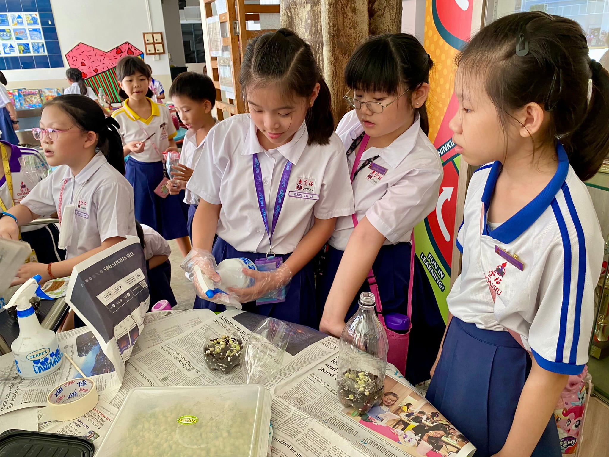 Young students in white uniforms work at a table covered with newspaper, handling small plants in bottles.