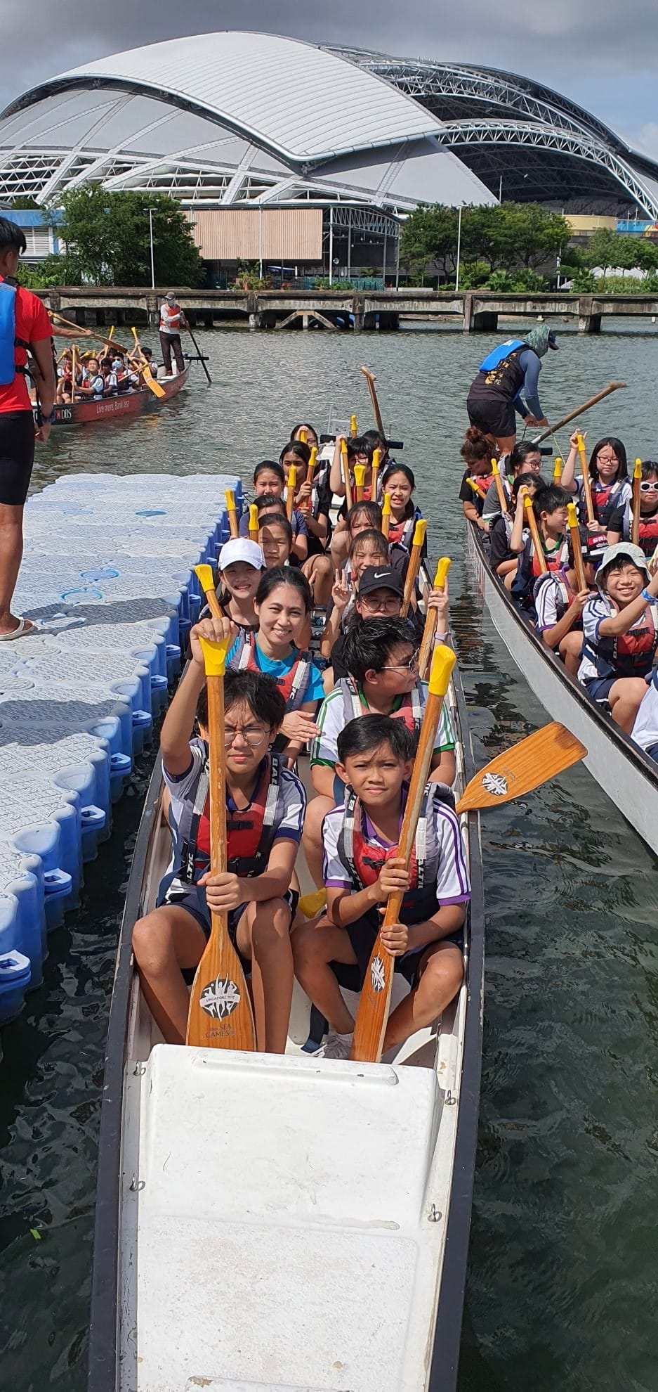 A group of children and adults holding paddles sit in a dragon boat on water near a large domed stadium.