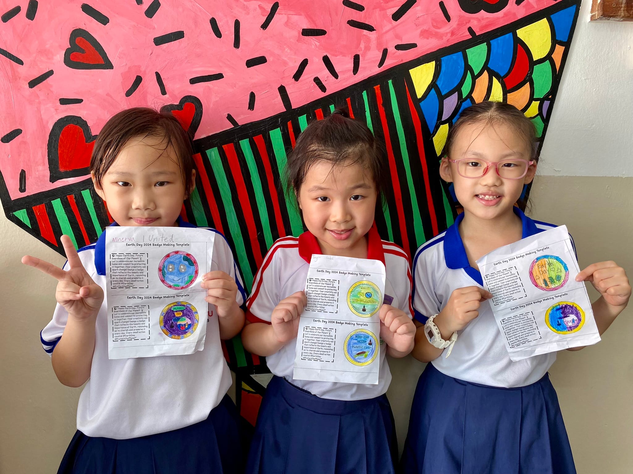Three young girls in school uniforms hold Earth Day 2024 badge-making templates in front of a colorful painted mural.