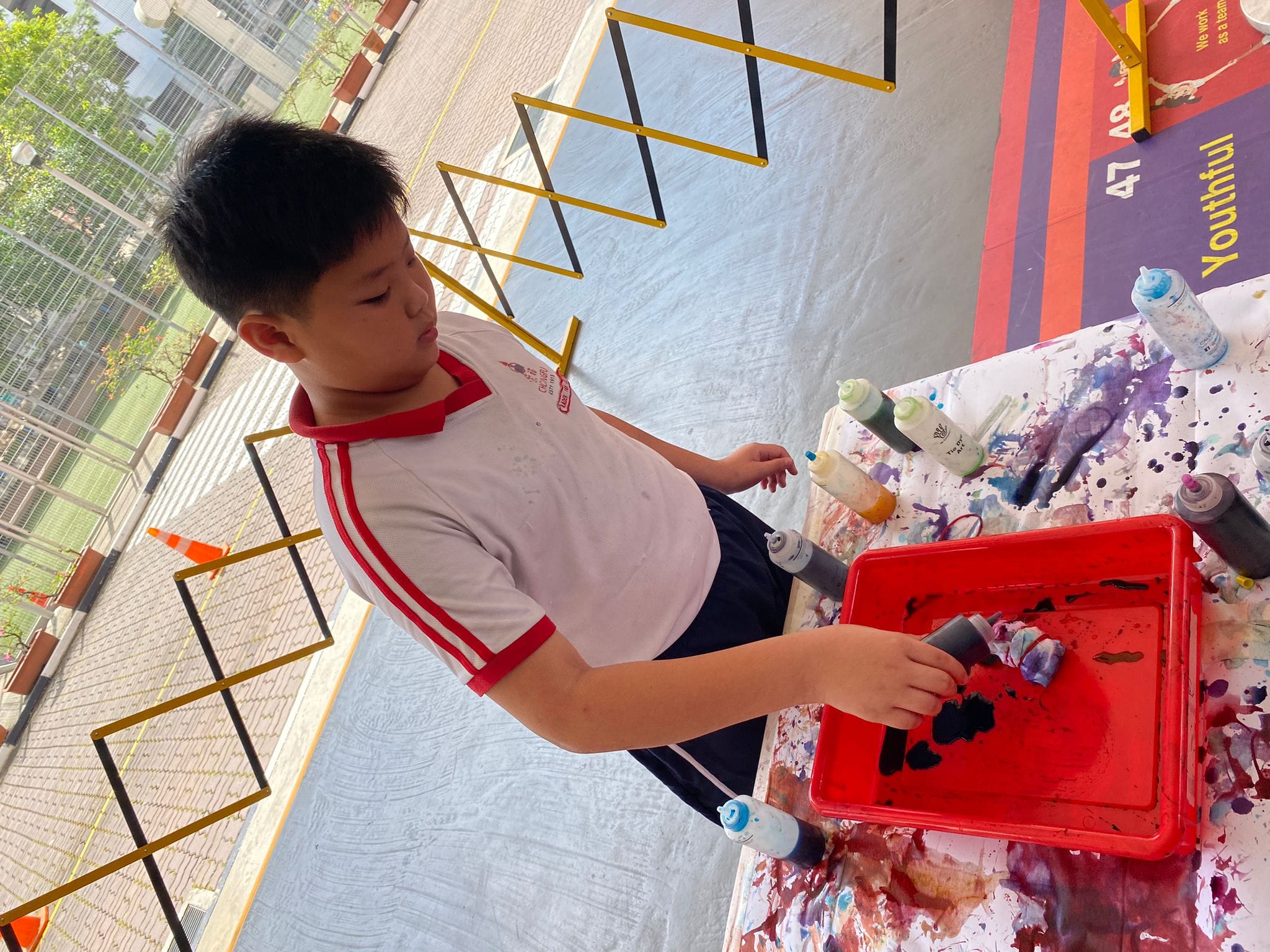 A young boy in a white and red shirt squirts paint into a red tray during an outdoor art activity.