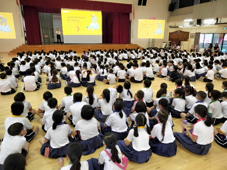 Students in white uniforms sit on a hall floor facing a stage with a yellow "Learn to Think by Seeing" presentation.