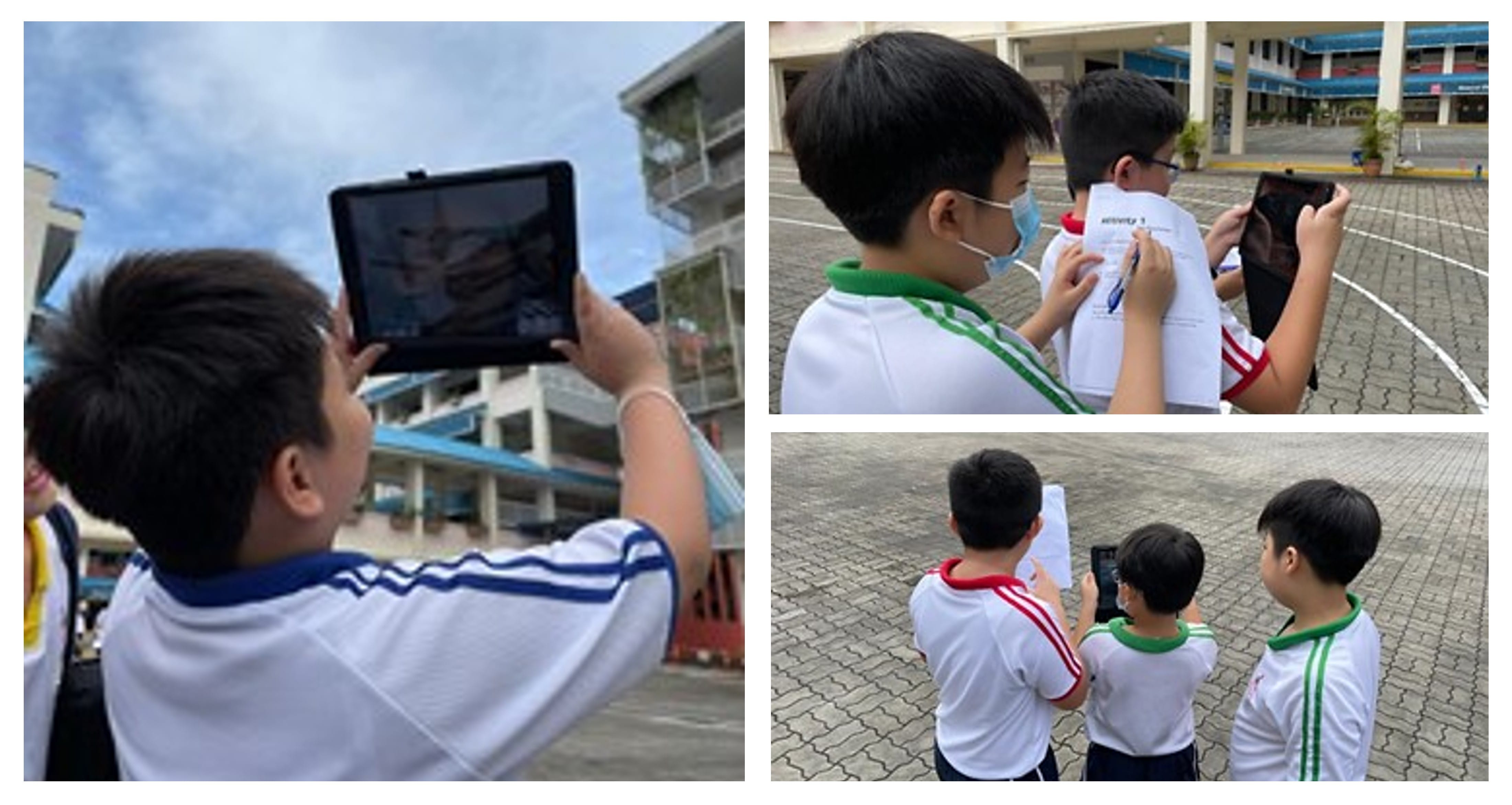 School boys in white uniforms use tablets and write on worksheets outdoors in a paved school courtyard.