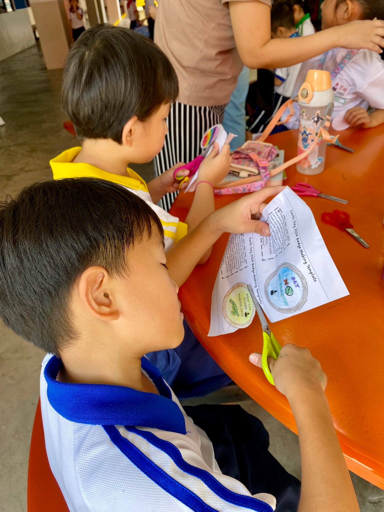 Two children in school uniforms cut Earth Day badge templates with scissors at an orange table.