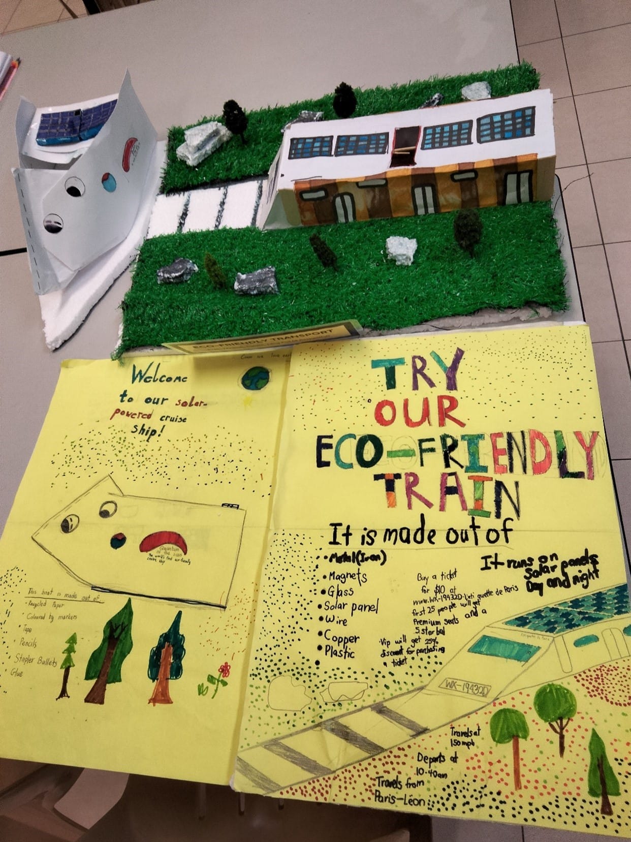 Student project displaying a handmade eco-friendly train poster and a cardboard building model on artificial grass.