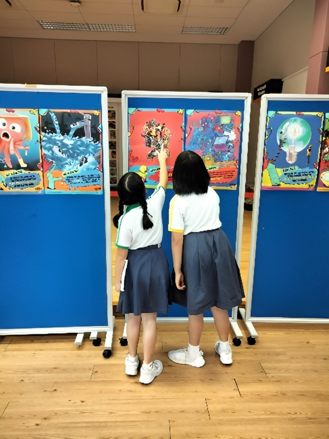 Two girls in school uniforms stand facing colorful artwork displayed on blue rolling boards indoors.