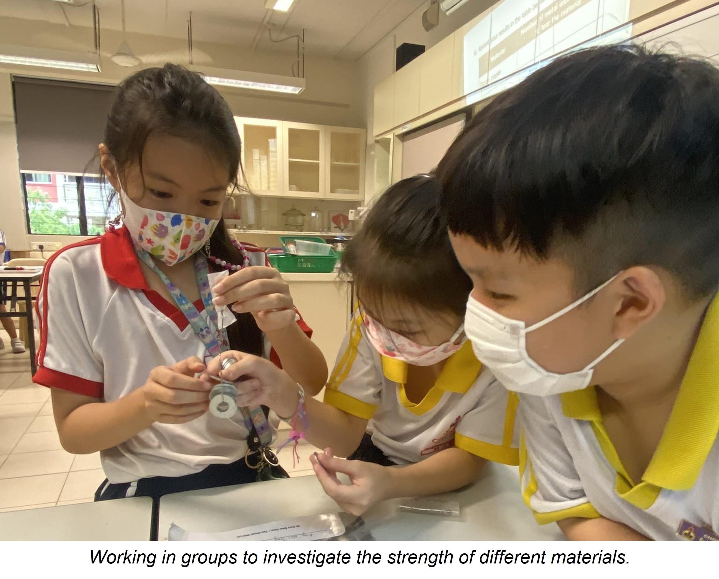 Three masked school children in uniforms work together at a desk, handling small objects in a classroom.