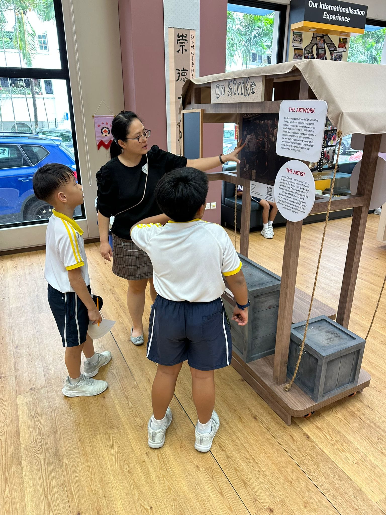 A woman and two young students in school uniforms examine an "On Strike" artwork display inside a school exhibition.