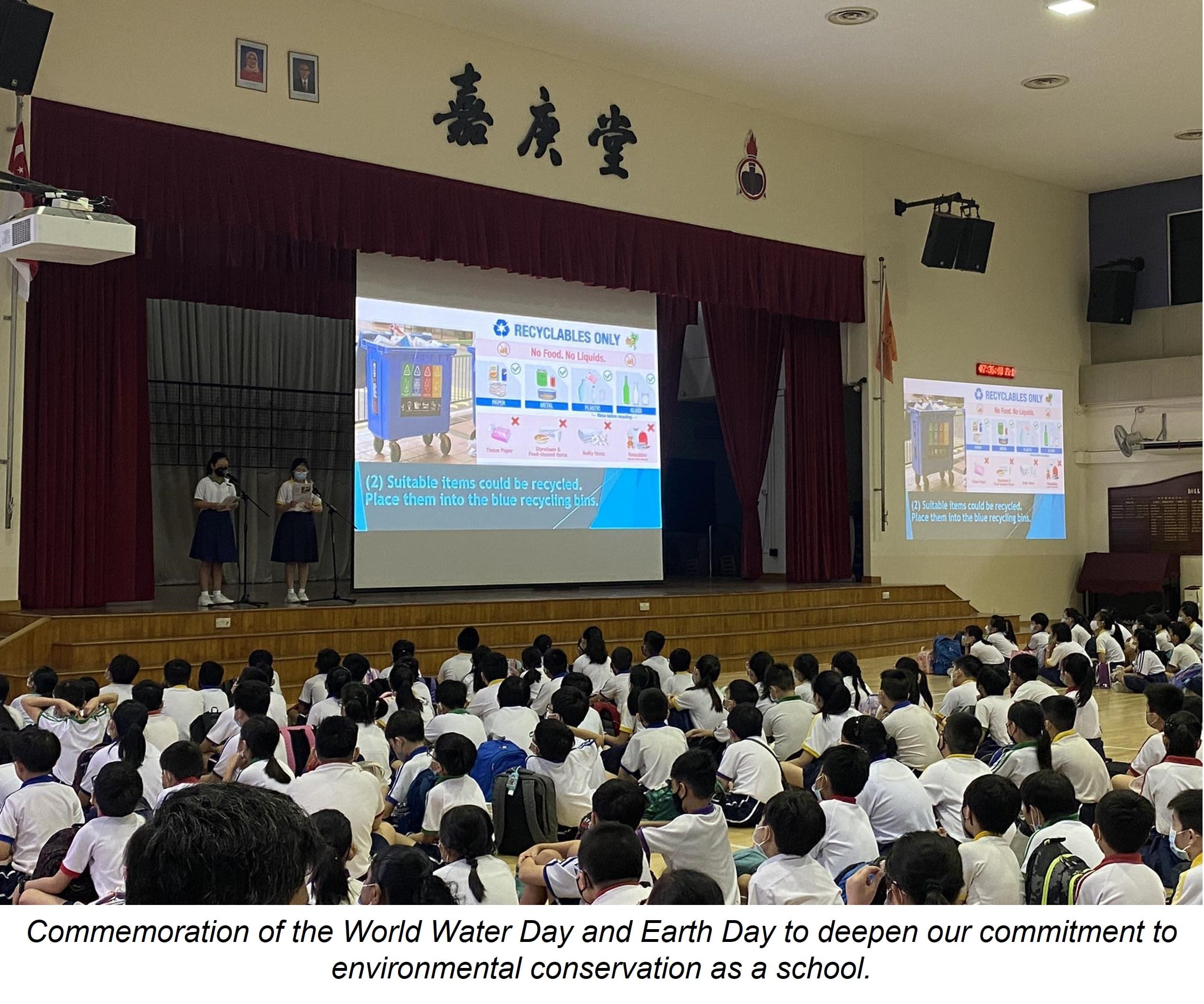 Students in a school hall watch a recycling presentation on stage showing a "Recyclables Only" slide.