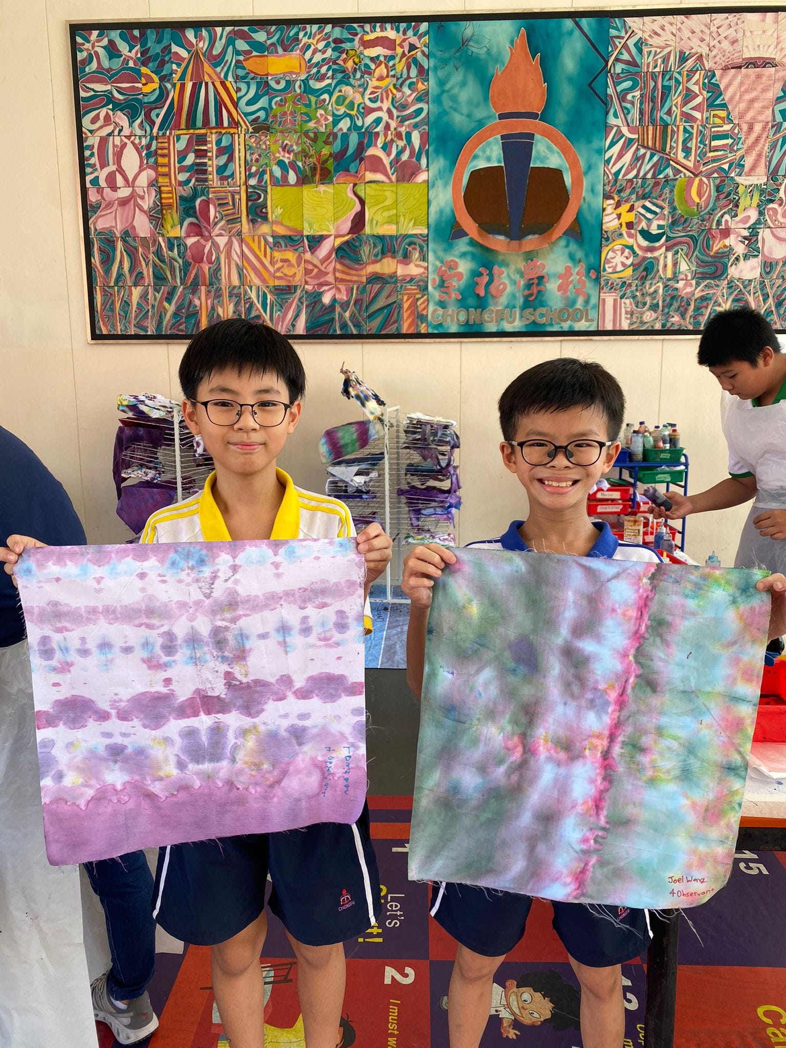 Two boys wearing glasses hold up colorful tie-dye fabric pieces inside Chongfu School.