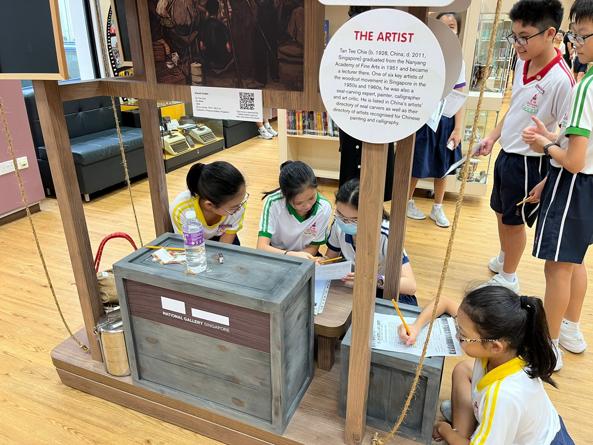 Students in school uniforms write notes at a National Gallery Singapore exhibit with an artist information sign.