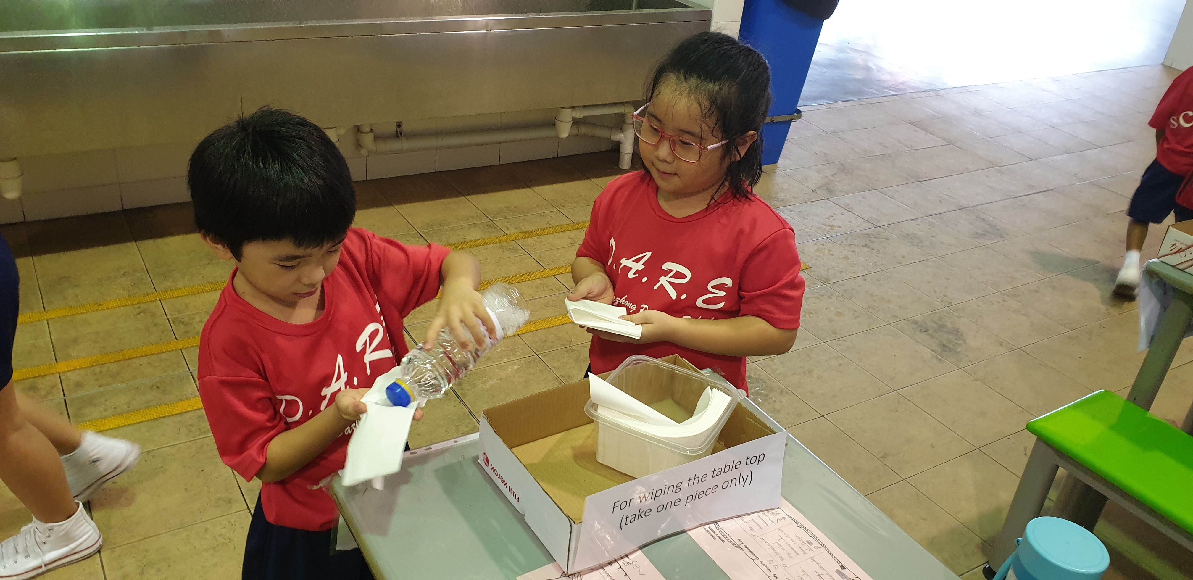 Two children in red D.A.R.E. shirts clean a table, one pouring water and the other holding a paper towel.