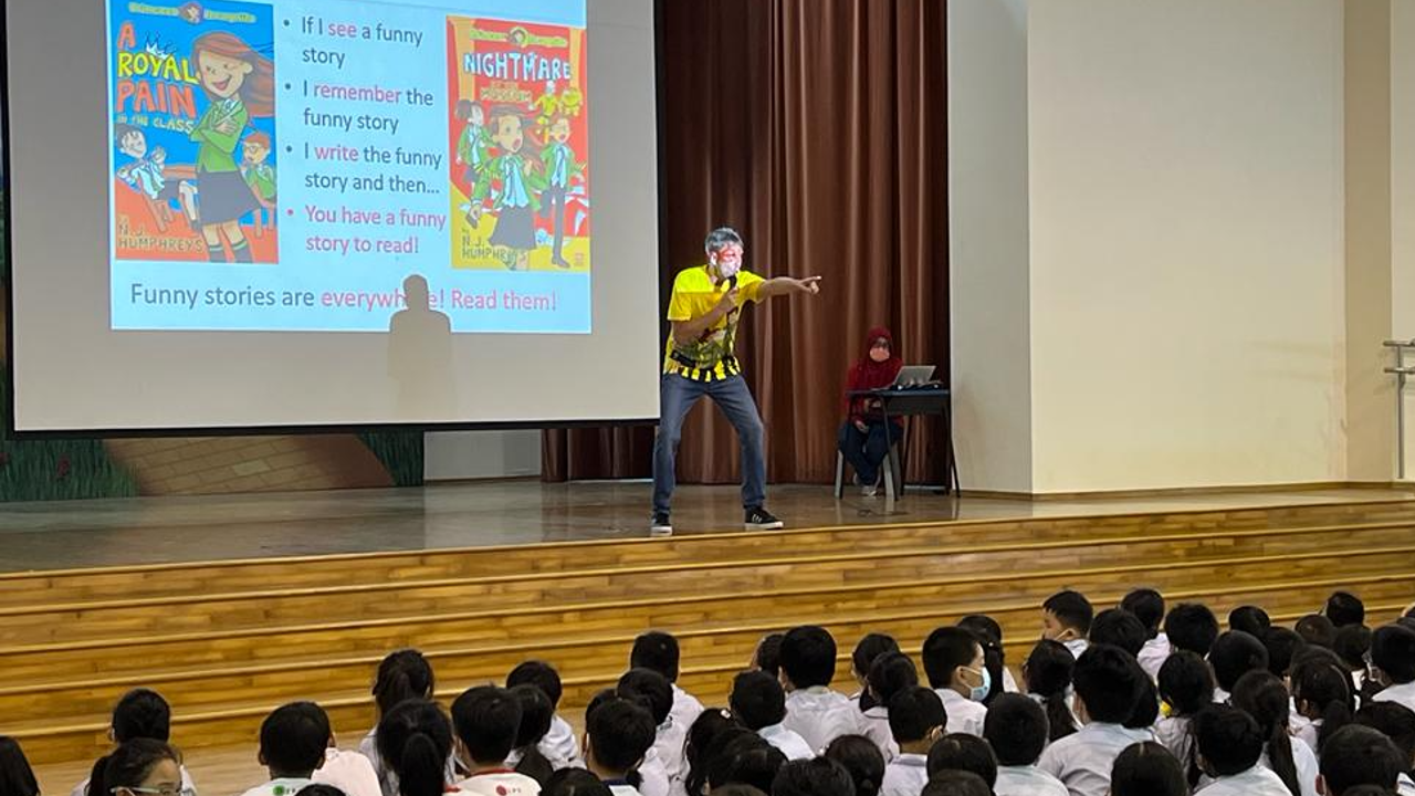 A presenter with face paint points at a school audience during a reading assembly with a slideshow about funny stories.